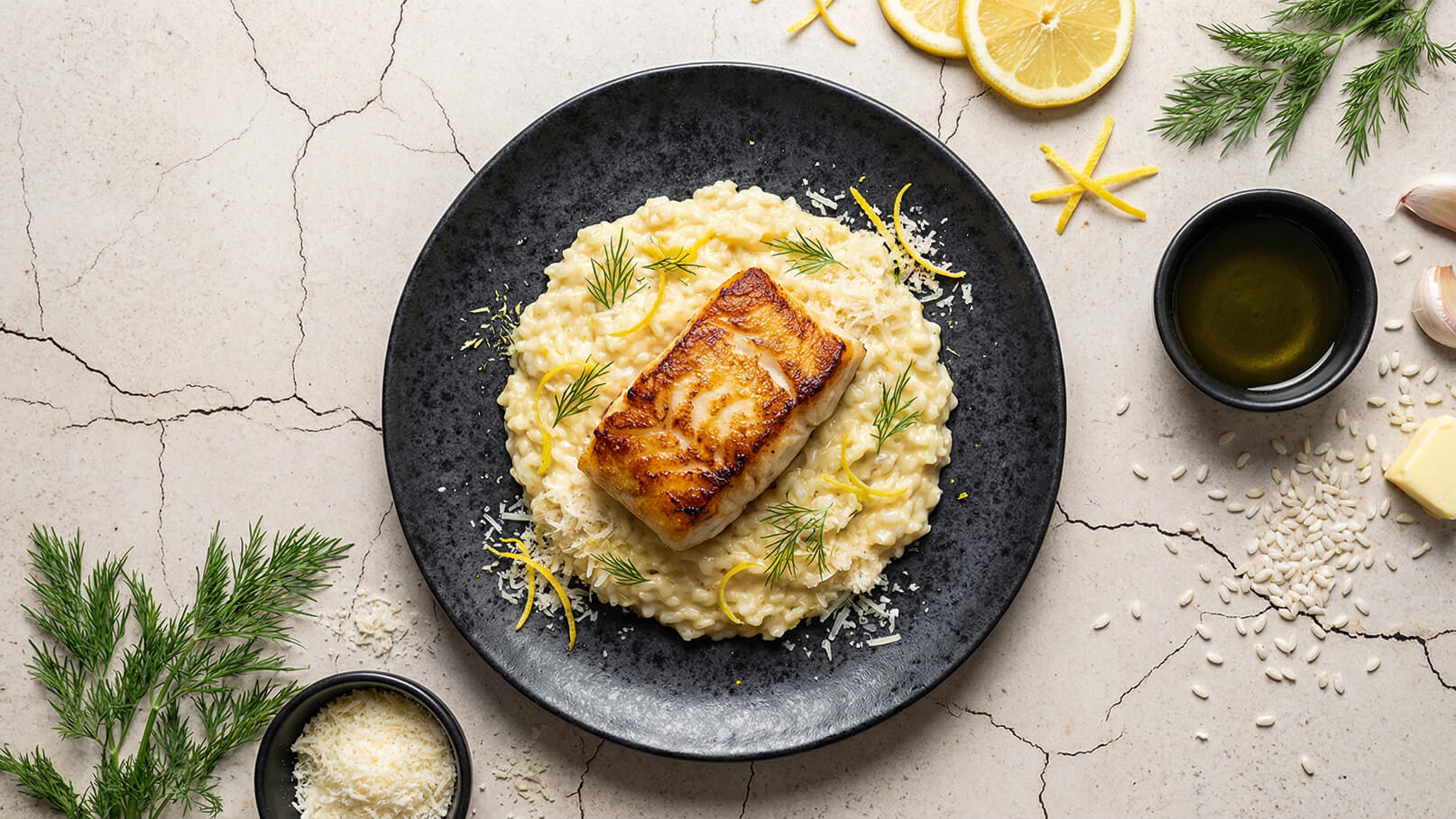 Ein dunkler Teller mit cremigem Risotto und einem Stück gebratenem Fisch. Das Gericht ist mit frischem Dill und Zitronenscheiben garniert. Neben dem Teller befinden sich einige Zutaten wie Käse, Olivenöl und Gewürze.