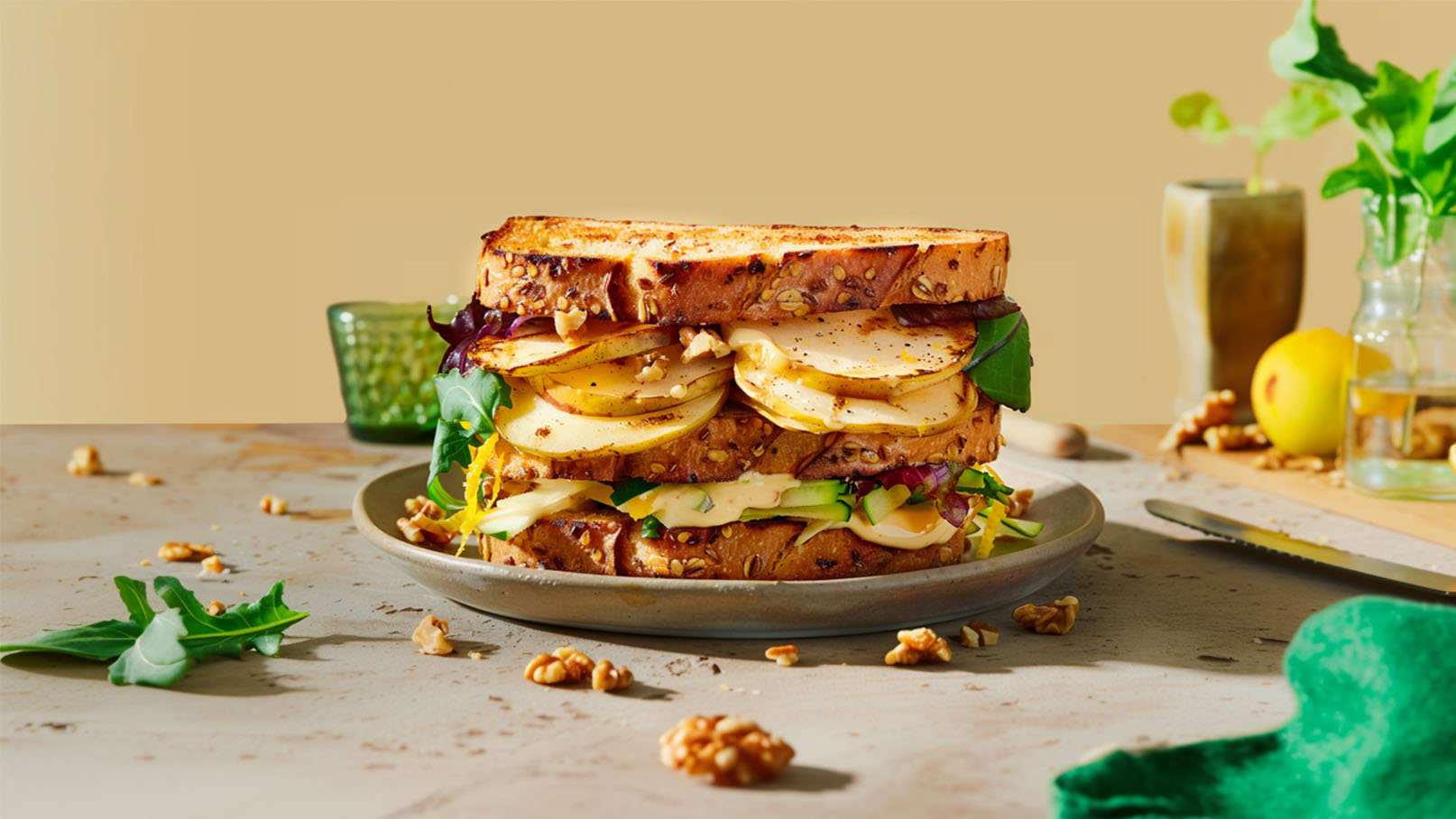 Ein üppig belegtes Sandwich auf einem grauen Teller. Es besteht aus mehreren Schichten Vollkornbrot, gefüllt mit Äpfeln, grünem Blattgemüse, Käse und anderen frischen Zutaten. Der Hintergrund ist einfarbig beige, und im Bild sind einige Nüsse und andere Speisen zu sehen.