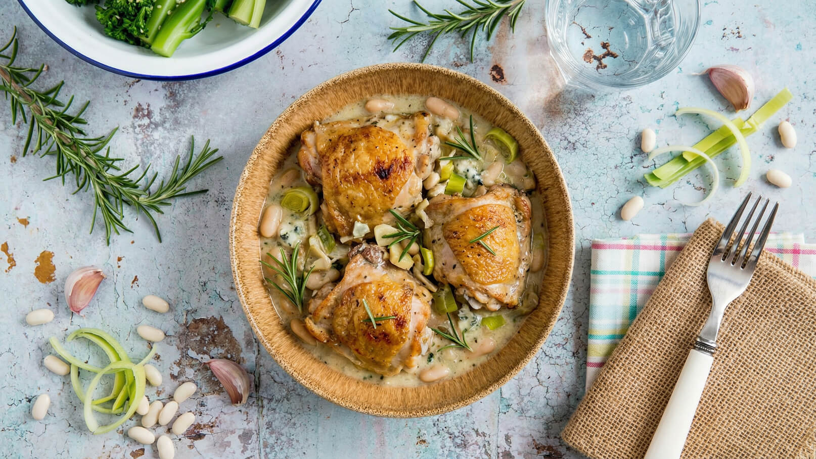 Ein Schalengericht mit zartem, gebratenem Hähnchen, umgeben von weißen Bohnen und Lauch in einer cremigen Sauce. Frischer Rosmarin verleiht dem Gericht Aroma. Im Hintergrund sind ein Glas Wasser, ein Stück Knoblauch und etwas geschnittenes Gemüse zu sehen.