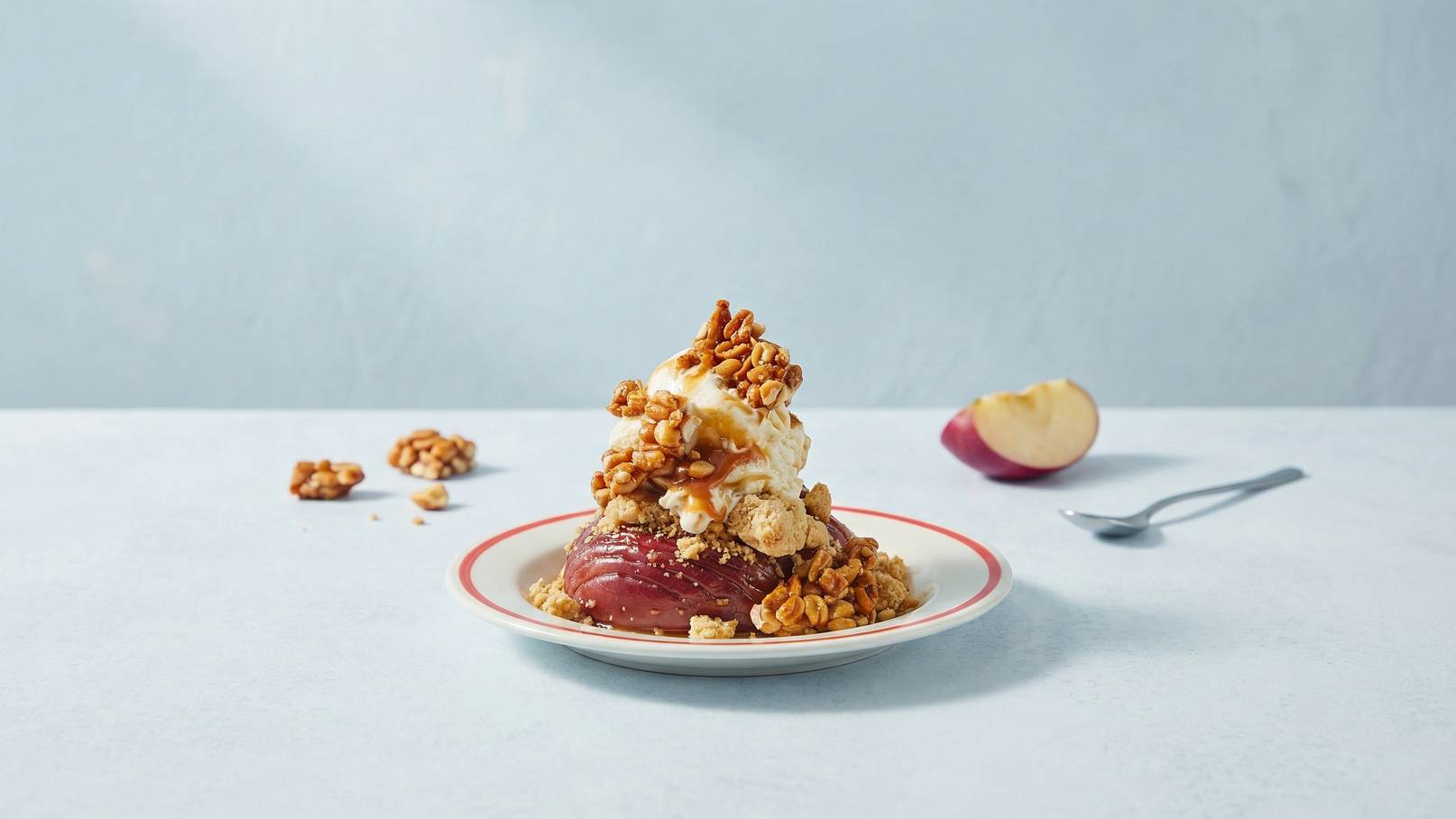 Eine Schüssel mit warmem, karamellisiertem Apfel, bedeckt mit einer Kugel Vanilleeis, knusprigen Karamellstücken und einer Prise Keksbrösel. Daneben liegen ein halber Apfel und einige Keksstücke auf einem blauen Tisch.