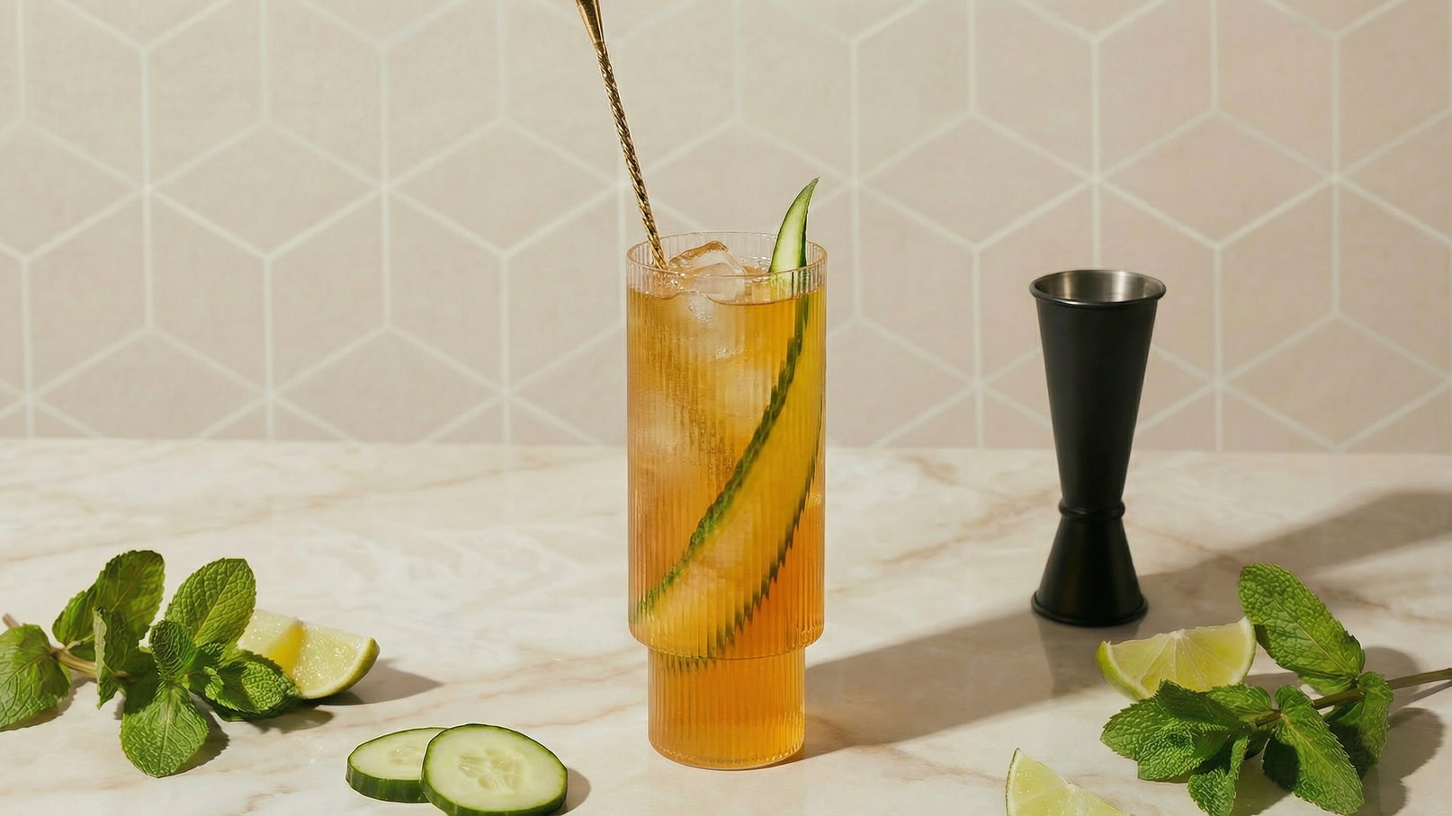 Ein hoher Cocktailglas mit einem erfrischenden Getränk, versehen mit Eiswürfeln und einer Gurkenscheibe, steht auf einem marmorierten Tisch. Neben dem Glas liegen frische Minzblätter, Limettenscheiben und einige Gurkenscheiben, während ein Messbecher aus schwarzem Metall daneben platziert ist.