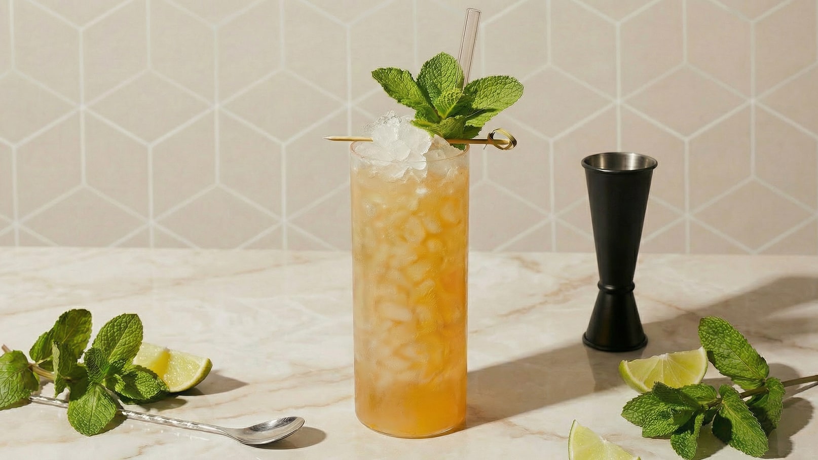 Ein Glas mit einem fruchtigen Cocktail, gefüllt mit Eisstücken und dekoriert mit frischen Minzblättern. Neben dem Glas liegen einige Limettenscheiben und ein Messbecher aus Metall auf einer marmorierten Oberfläche. Der Hintergrund ist in einem hellen geometrischen Muster.