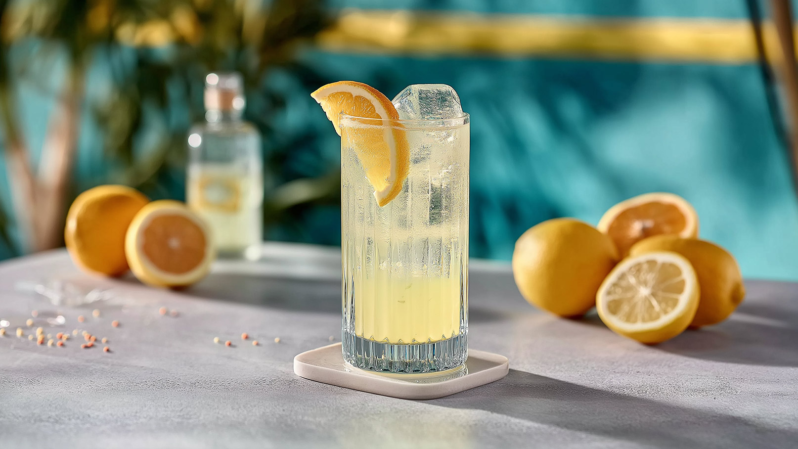 Ein erfrischendes Getränk in einem hohen Glas mit Eiswürfeln und einer Zitronenscheibe. Im Hintergrund liegen mehrere Zitronen und eine Flasche mit einer klaren Flüssigkeit. Der Tisch hat eine helle, graue Oberfläche.