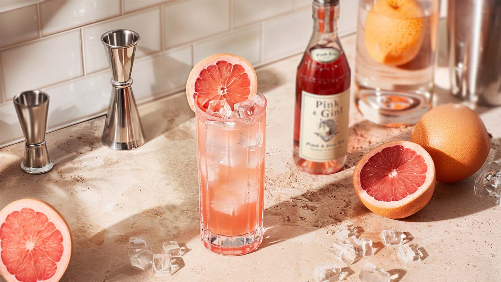 Ein Cocktailglas mit Eiswürfeln und einer Scheibe Grapefruit steht auf einer Tischoberfläche. Im Hintergrund sind eine Flasche Grapefruit-Getränk und eine halbierte Grapefruit zu sehen. Die Szene vermittelt einen erfrischenden, fruchtigen Eindruck.