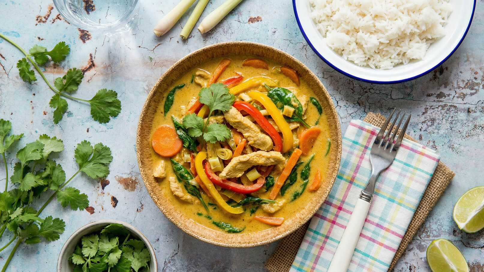 Ein Schüssel mit einer bunten Gemüsecurry-Mischung aus Hähnchenbrust, Karotten, Paprika und Spinat ist in der Mitte des Bildes zu sehen. Daneben befindet sich eine Schüssel mit Reis und frische Korianderblätter auf dem Tisch. Ein Besteckset ist ebenfalls vorhanden.