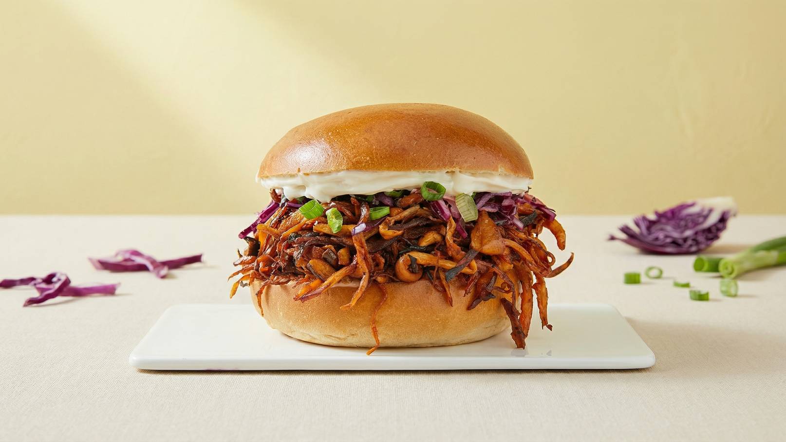 Ein Sandwich auf einem weißen Teller, gefüllt mit zerrupftem Fleisch und lila Kraut, garniert mit Frühlingszwiebeln und einer cremigen Sauce. Der Brötchenrand ist goldbraun und die Füllung sieht saftig und aromatisch aus. Der Hintergrund ist ein weiches Gelb.