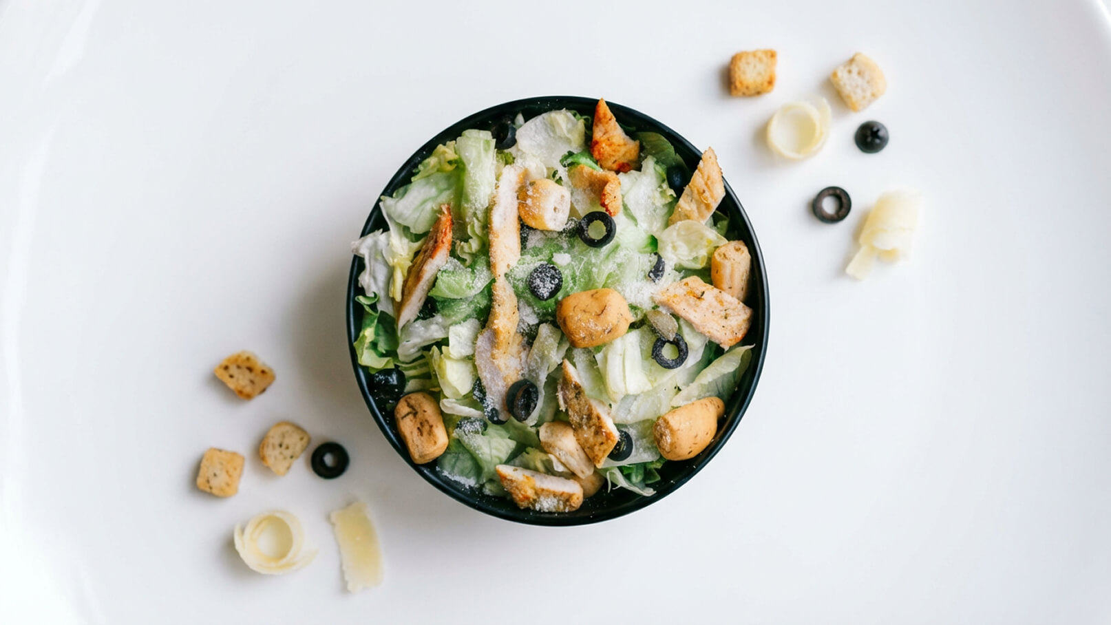 Eine schwarze Schüssel mit grünem Salat, Stückchen gegrilltem Hähnchen, schwarzen Oliven und Croutons. Der Salat sieht frisch aus und ist mit Parmesan bestreut. Um die Schüssel herum liegen einige weitere Croutons und Käsestreifen.