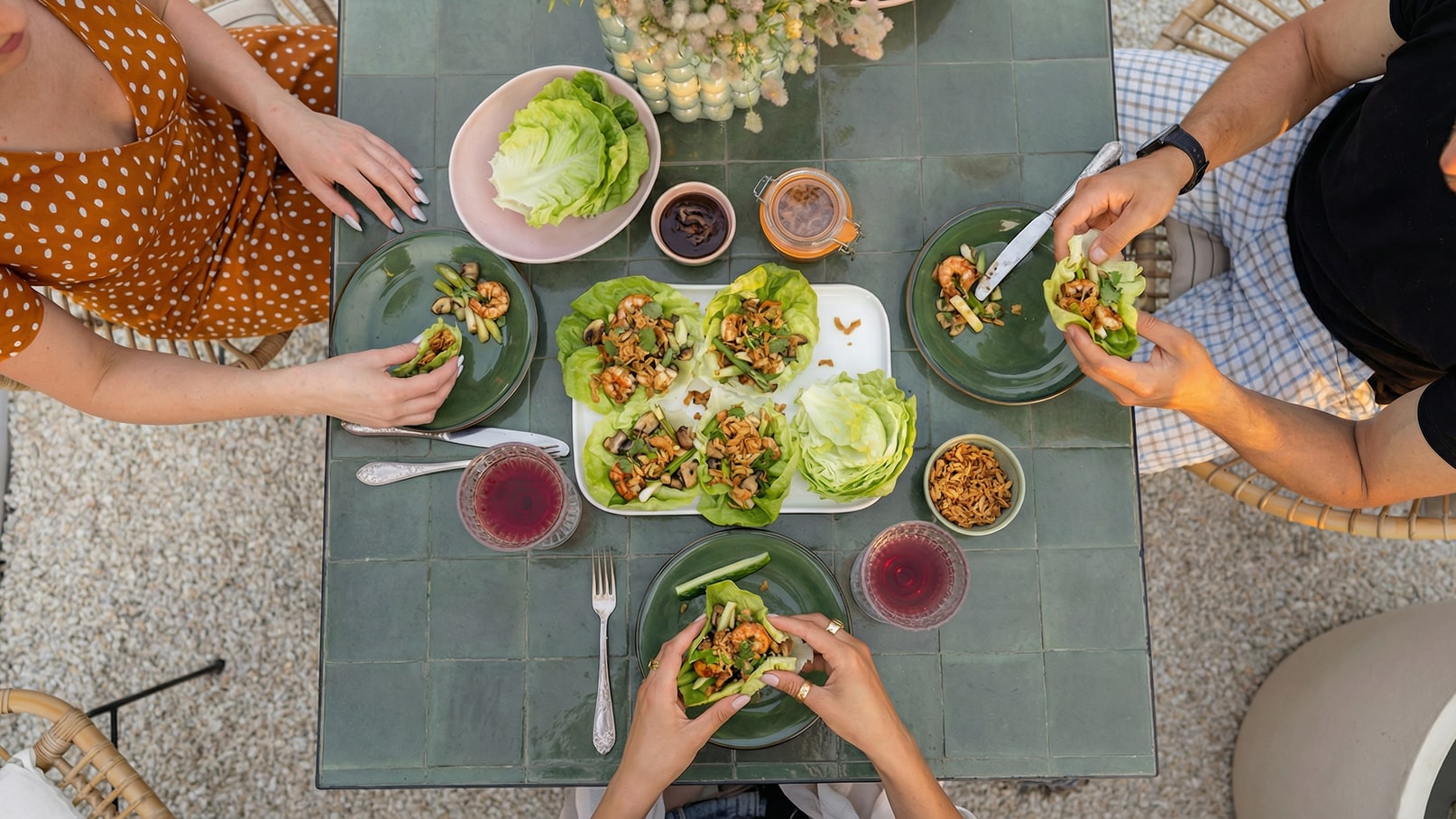 Auf einem Tisch sind verschiedene Teller mit Salat und einer Mischung aus Garnelen und anderen Zutaten angerichtet. Zwei Personen greifen nach Salatblättern, um ihre Speisen zuzubereiten. Rote Getränke und kleine Schalen mit weiteren Zutaten sind ebenfalls vorhanden.