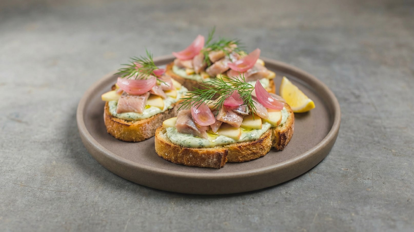 Drei kleine Brotscheiben liegen auf einem grauen Teller. Sie sind mit einer cremigen Sauce, Fischstücken, Äpfeln, roten Zwiebeln und frischem Dill garniert. Ein Stück Zitrone komplettiert die Präsentation.
