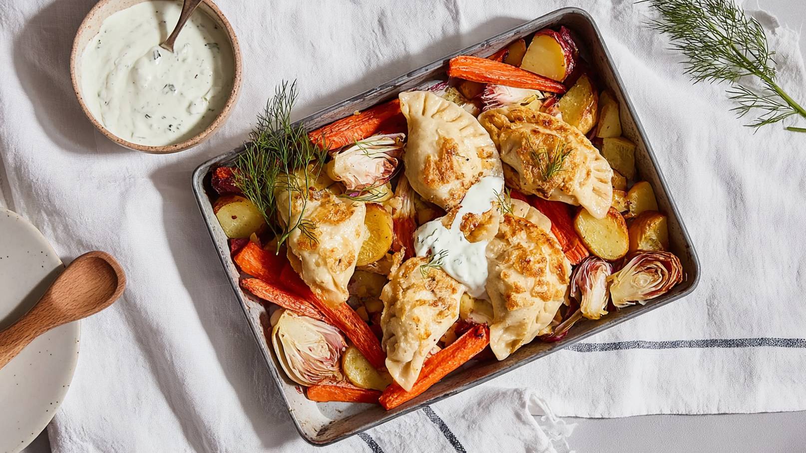 Ein Blech mit goldbraun gebratenen Teigtaschen liegt auf einem weißen Tisch. Umgeben von buntem, geröstetem Gemüse wie Karotten und Kartoffeln. Neben dem Blech steht eine Schüssel mit einer grünen, cremigen Soße.