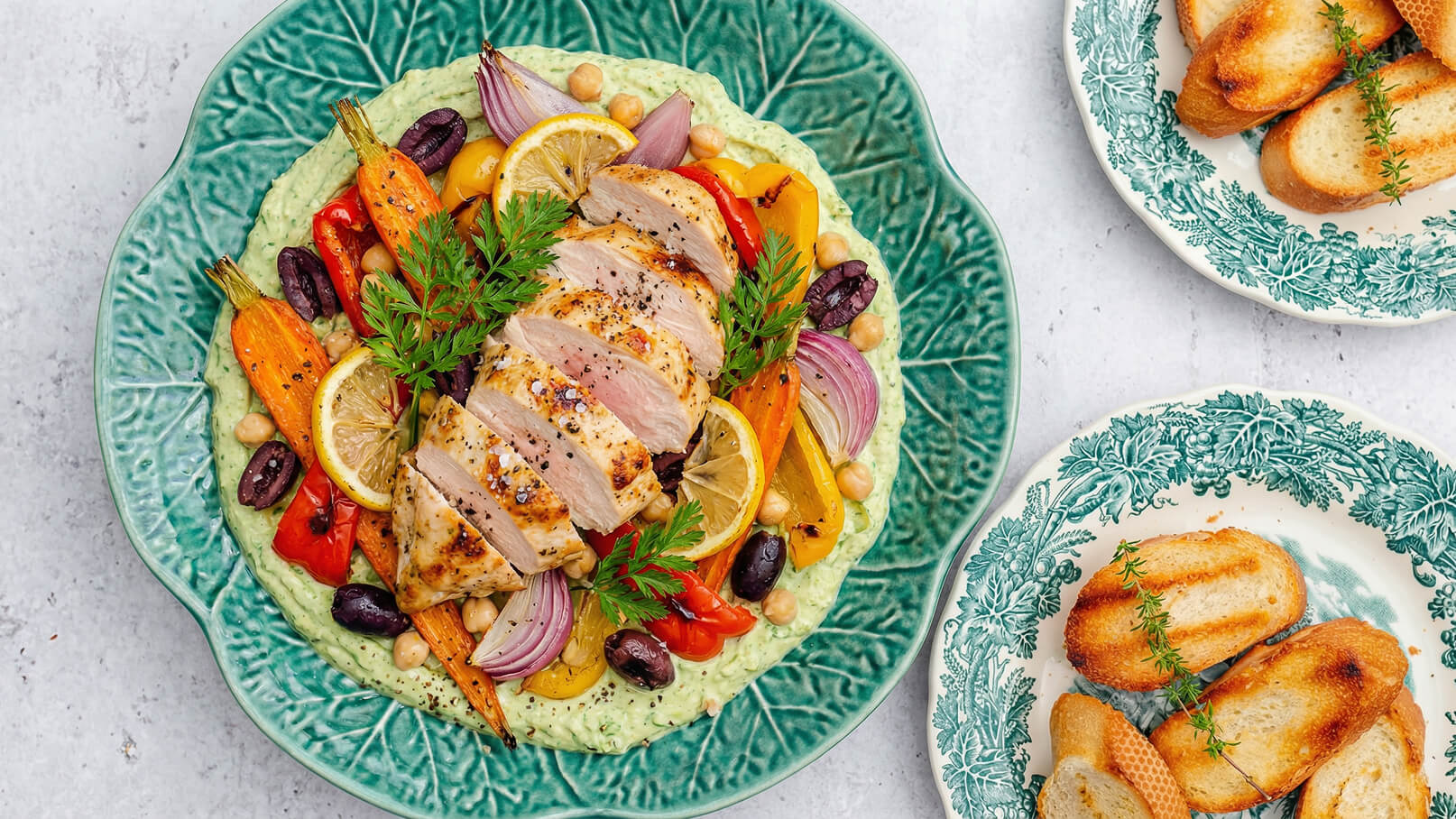 Ein grüner Teller mit püriertem Gemüse als Basis, garniert mit gegrilltem Hühnchen, buntem Gemüse wie Karotten, Paprika und roten Zwiebeln sowie Zitronenscheiben. Daneben befinden sich zwei weiße Teller mit geröstetem Brot.