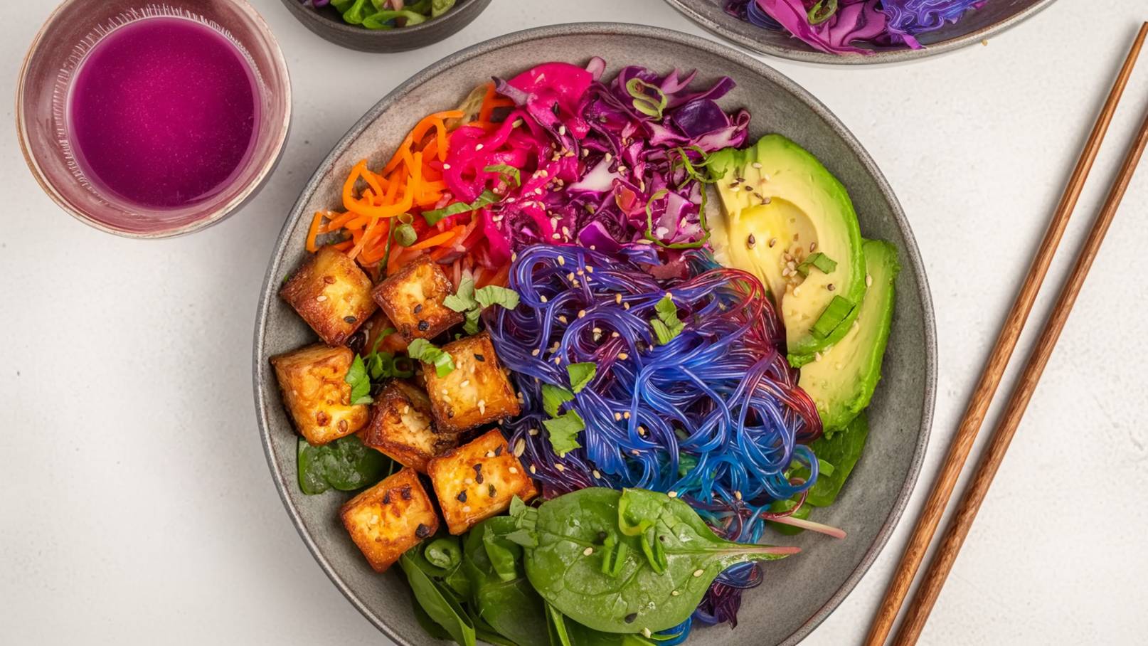 Eine Schüssel mit bunten Nudeln liegt im Vordergrund, umgeben von gebratenem Tofu, frischem Spinat, Karottenstreifen, Rotkohl und Avocado. Im Hintergrund sind einige kleine Schalen mit weiteren Zutaten und ein Glas mit einem pinken Getränk zu sehen.
