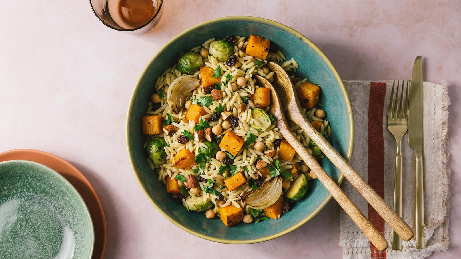 Eine große Schüssel mit Orzo-Pasta, Kürbiswürfeln, Rosenkohl, Kichererbsen, Mandeln und frischer Petersilie ist dekorativ angerichtet. Zwei Holzlöffel liegen neben dem Gericht. Im Hintergrund sind zwei kleine Teller und Besteck zu sehen.