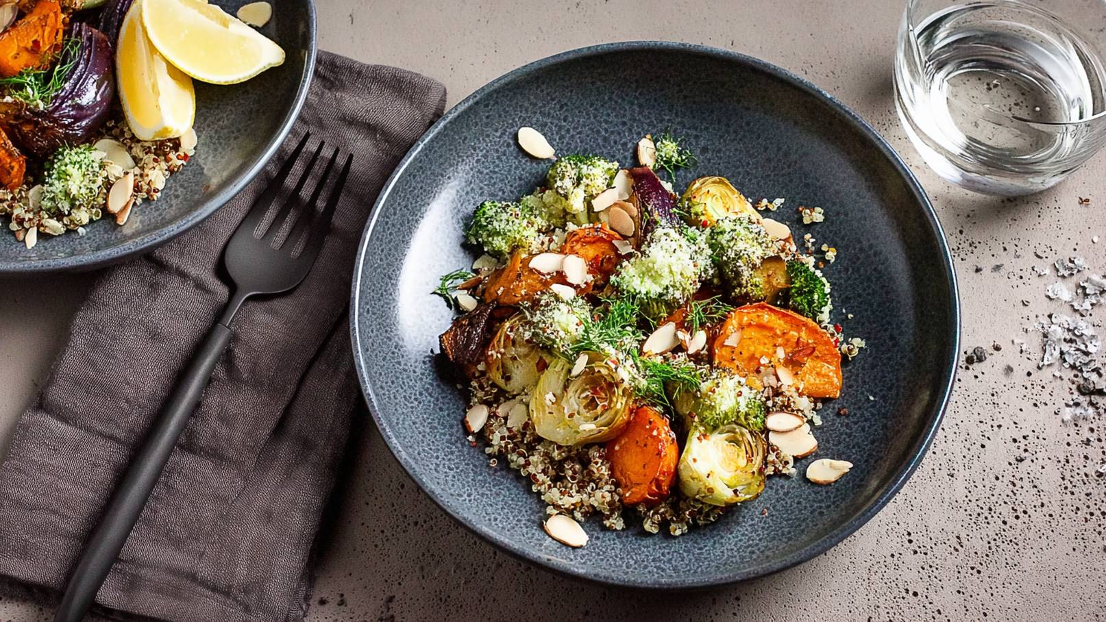 Eine dunkle Schale mit einer bunten Mischung aus geröstetem Gemüse, darunter Karotten, Brokkoli und Zwiebeln, ist auf einem Tisch angerichtet. Darunter liegt eine Schicht Quinoa, garniert mit Mandelsplittern und frischem Dill. Neben der Schale befindet sich eine Gabel und ein Wasserglas.