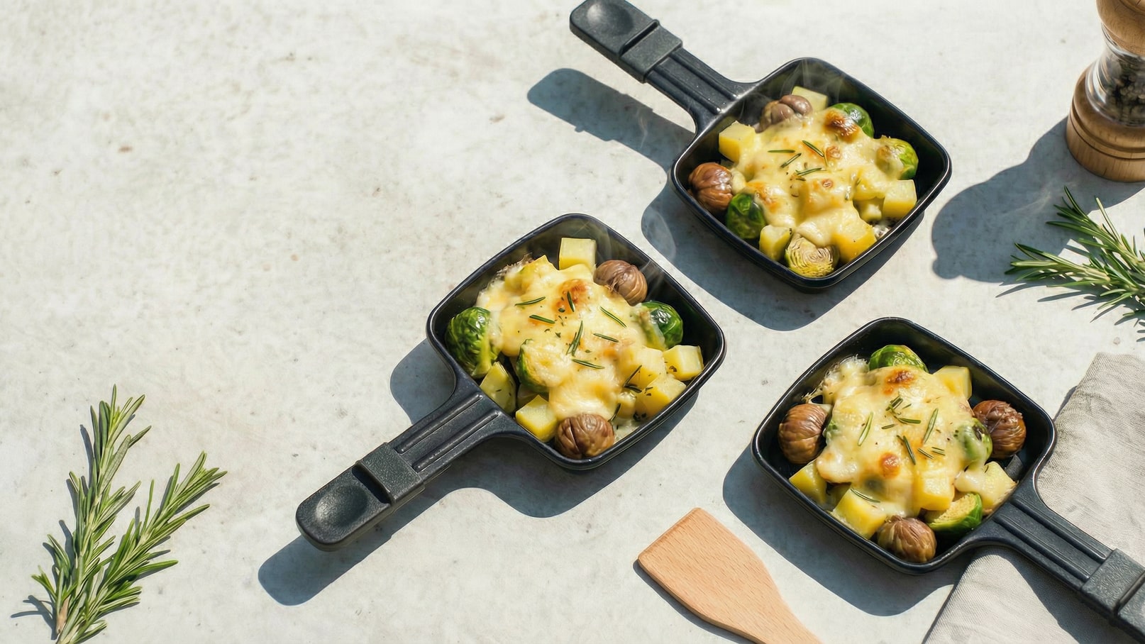 Drei kleine Pfannen mit geschmolzenem Käse, Kartoffeln, Rosenkohl und Kastanien sind auf einem hellen Untergrund platziert. Die Pfannen sind schwarz mit einem Griff und das Essen wird von frischen Kräutern begleitet.