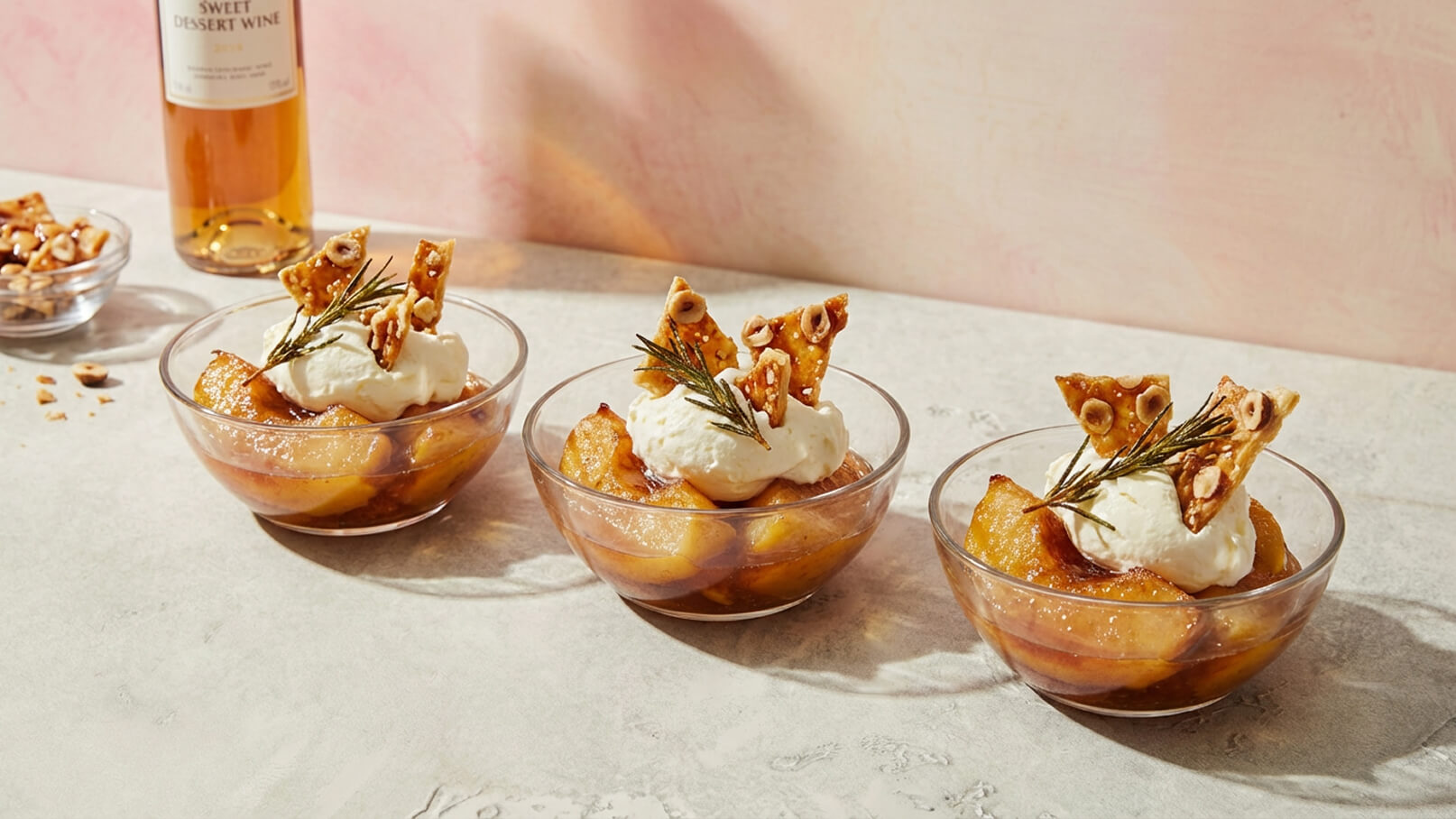 Drei Schalen mit karamellisierten Äpfeln, die mit Sahne und knusprigem Gebäck dekoriert sind, stehen auf einem Tisch. Einige Nüsse sind in einer separaten Schale daneben. Eine Flasche süßer Dessertwein ist im Hintergrund zu sehen.