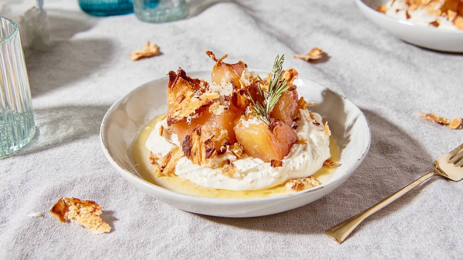 Eine Schüssel mit einer cremigen Dessertbasis aus Sahne, die mit karamellisierten Äpfeln und knusprigen Stücken bedeckt ist. Ein Zweig Rosmarin dekoriert das Gericht. Im Hintergrund sind weitere Tellerschalen und kleine Gläser sichtbar.