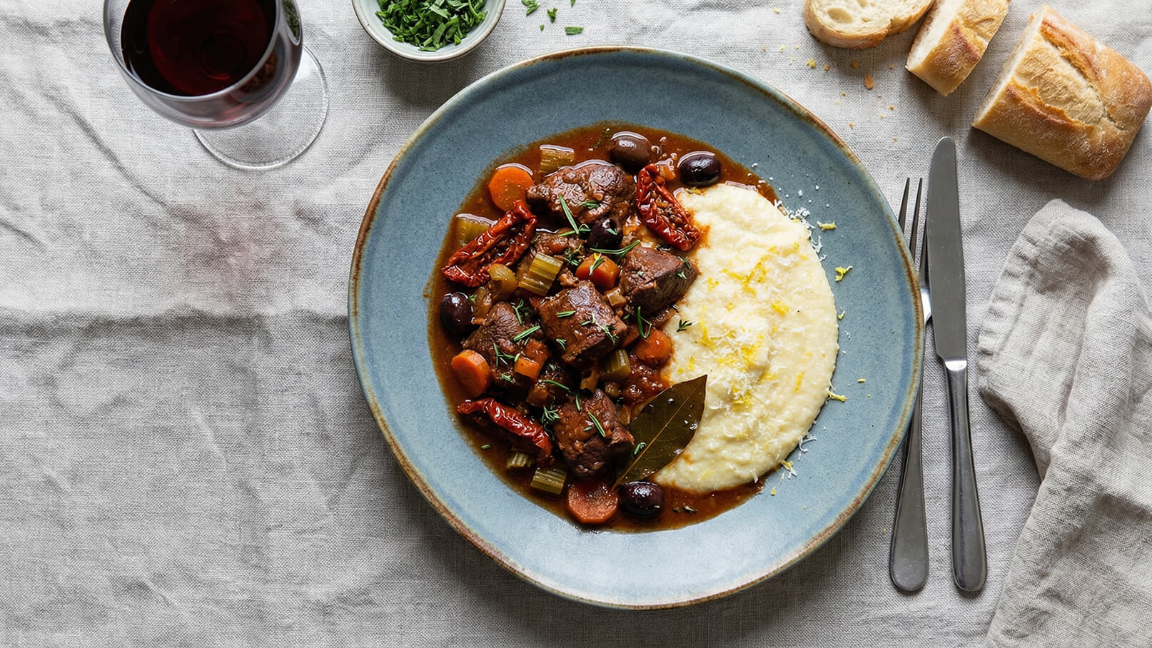 Ein Teller mit aufwendig zubereitetem Fleischgericht in einer richartigen Soße, garniert mit frischen Kräutern und verschiedenen Gemüsesorten. Daneben befindet sich eine Portion cremiger Polenta. Serviert wird dazu ein Glas Rotwein. Im Hintergrund ist ein Stück Brot sichtbar.