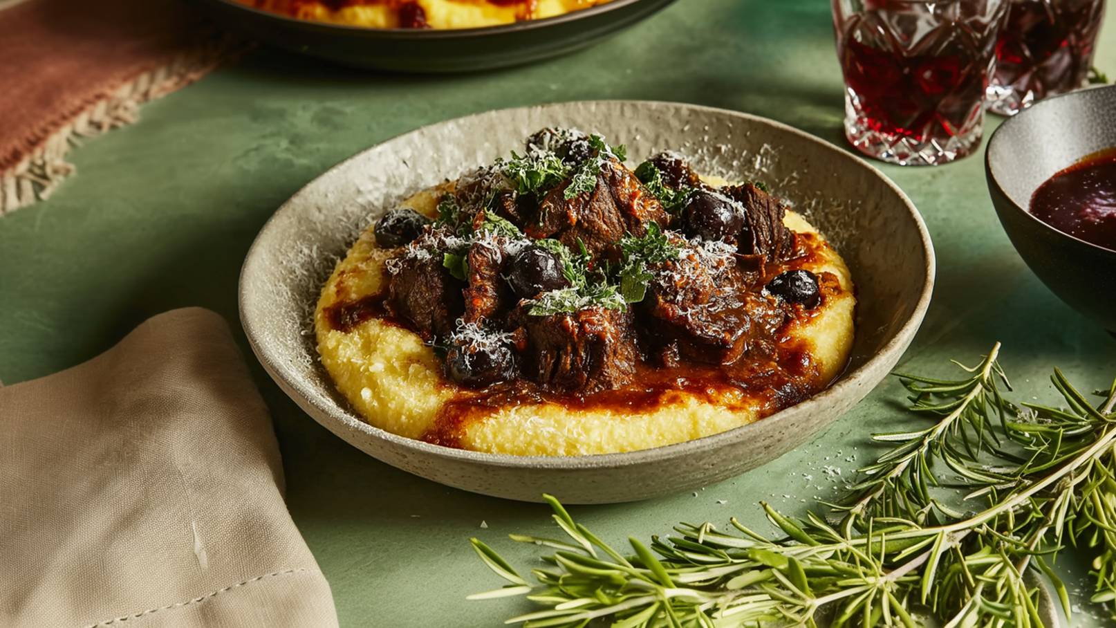 Ein Teller mit cremigem Polenta, bedeckt mit zartem, geschmortem Fleisch und schwarzen Oliven. Das Gericht ist mit frischen Kräutern und geriebenem Käse garniert. Im Hintergrund sind Getränke und ein Rosmarinzweig zu sehen.