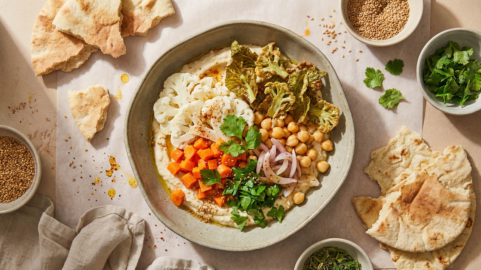 Eine Schüssel mit Hummus, garniert mit Blumenkohl, Karotten, Kichererbsen, Zwiebeln und frischen Kräutern. Daneben liegen einige Stücke Fladenbrot und Gewürze. Die Präsentation ist farbenfroh und appetitlich.