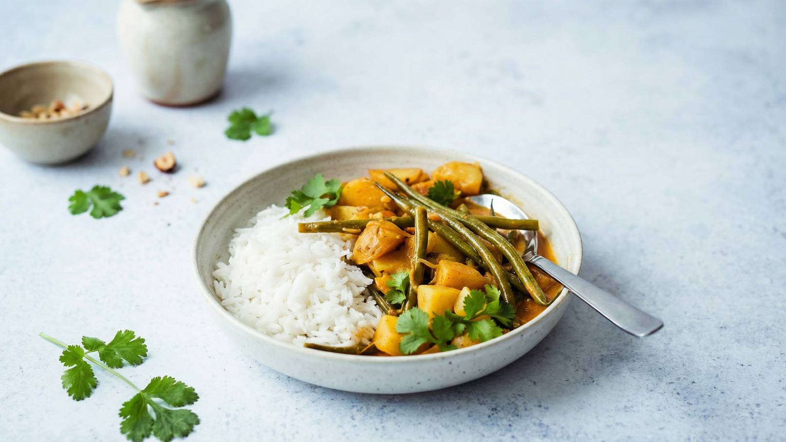 Ein teller mit weißem Reis und einem bunten Curry aus Kartoffeln, grünen Bohnen und Koriander. Der Teller steht auf einem blauen Untergrund, daneben befinden sich einige Korianderblätter und eine kleine Schüssel mit Nüssen.