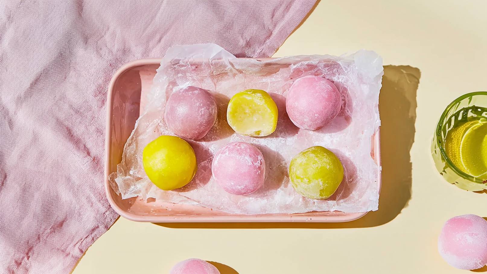 Auf einem rosa Teller liegen sechs runde, gefrorene Mochi. Die Mochi sind in verschiedenen Farben: pink, gelb und grün. Sie sind auf einem Stück durchsichtiger Folie platziert, umgeben von einem hellen, einfarbigen Hintergrund.
