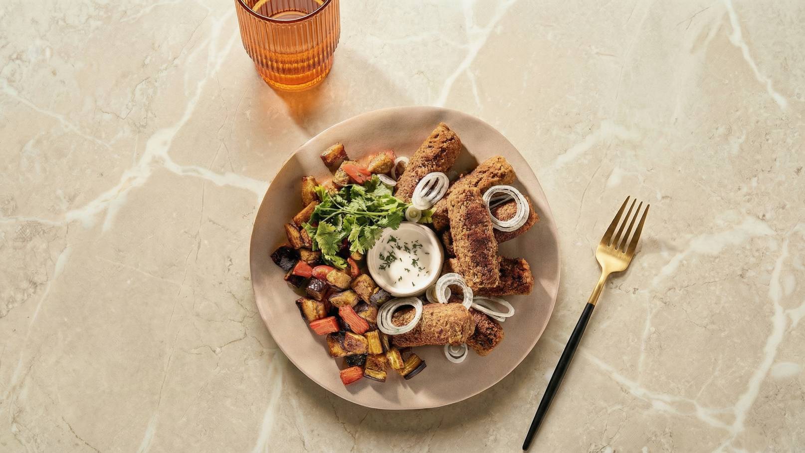 Ein Teller mit knusprigen, frittierten Rollen, garniert mit Zwiebelringen und frischem Koriander. Nebenbei ist ein Dip in einer kleinen Schale und eine bunte Mischung aus gebratenem Gemüse wie Auberginen, Paprika und Karotten. Ein goldener Besteck liegt daneben.