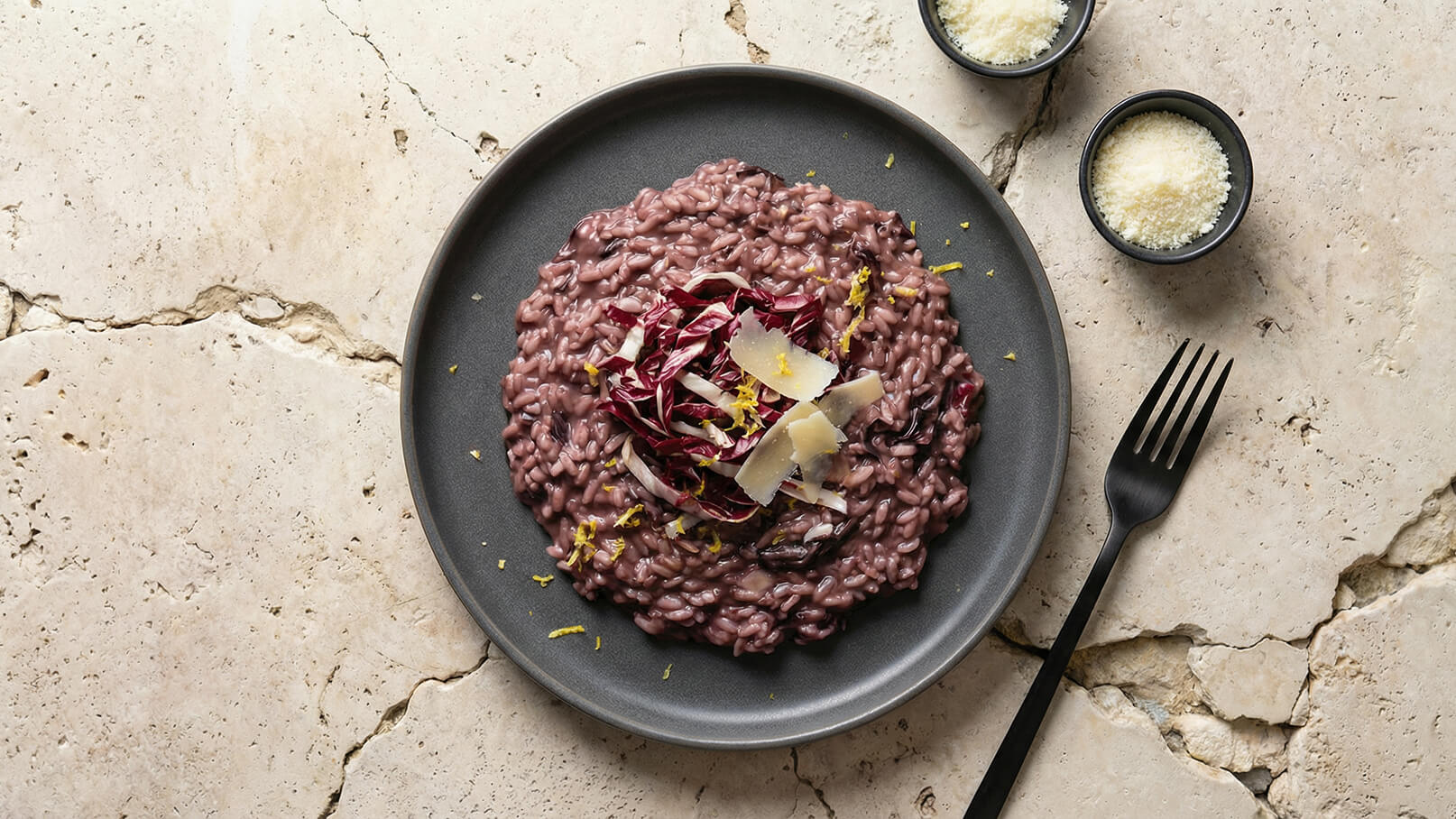 Ein Teller mit rosa Risotto, garniert mit roten Salatstreifen und Parmesanstücken, liegt auf einer neutralen, hellen Oberfläche. Daneben zwei Schalen mit geriebenem Parmesan und eine schwarze Gabel.