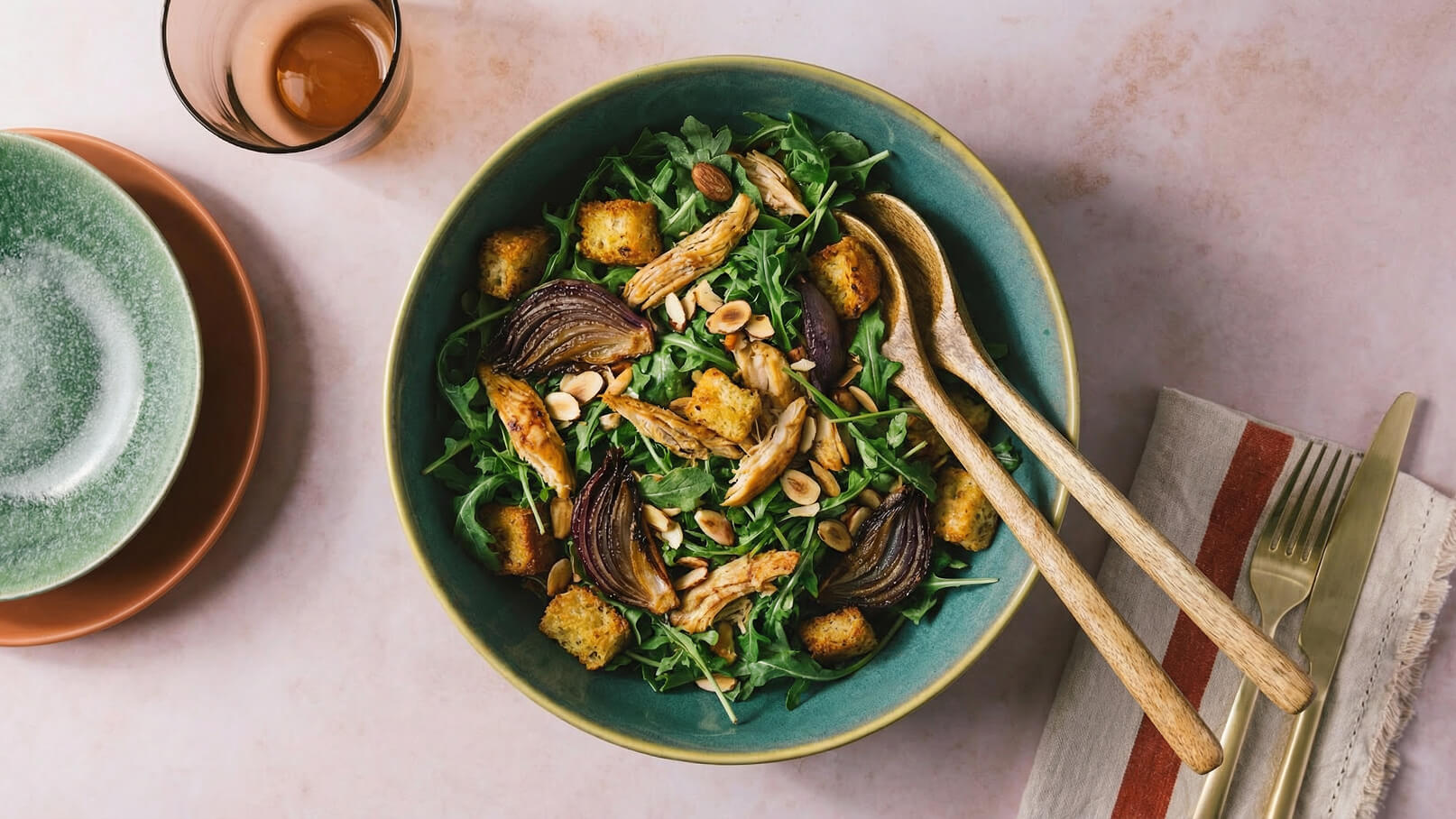 Eine große Schüssel Salat mit Rucola, gebratenen Zwiebeln, Hähnchenstücken und Croutons. Darüber sind geröstete Mandeln verteilt. Zwei Holzlöffel liegen in der Schüssel. Im Hintergrund befinden sich Geschirr und Besteck.