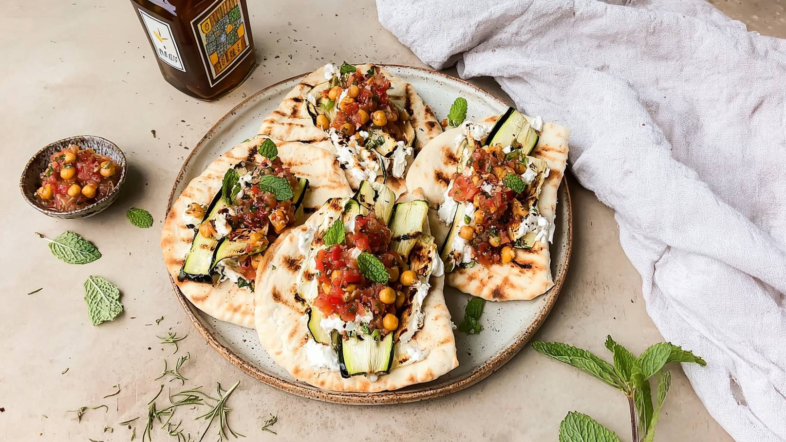 Auf einem Teller liegen gegrillte Fladenbrote, gefüllt mit Zucchini, Kräutern und einer bunten Salsa aus Tomaten und Kichererbsen. Frische Minzblätter dekorieren das Gericht. Neben dem Teller befindet sich eine kleine Schale mit zusätzlicher Salsa.