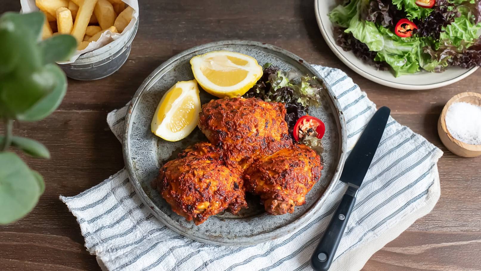 Ein Teller mit drei knusprigen, gewürzten Hähnchenteilen, serviert mit Zitronenscheiben und frischem Salat. Daneben liegt ein Besteckmesser auf einem gestreiften Tuch. Im Hintergrund befindet sich eine Schale mit Pommes.