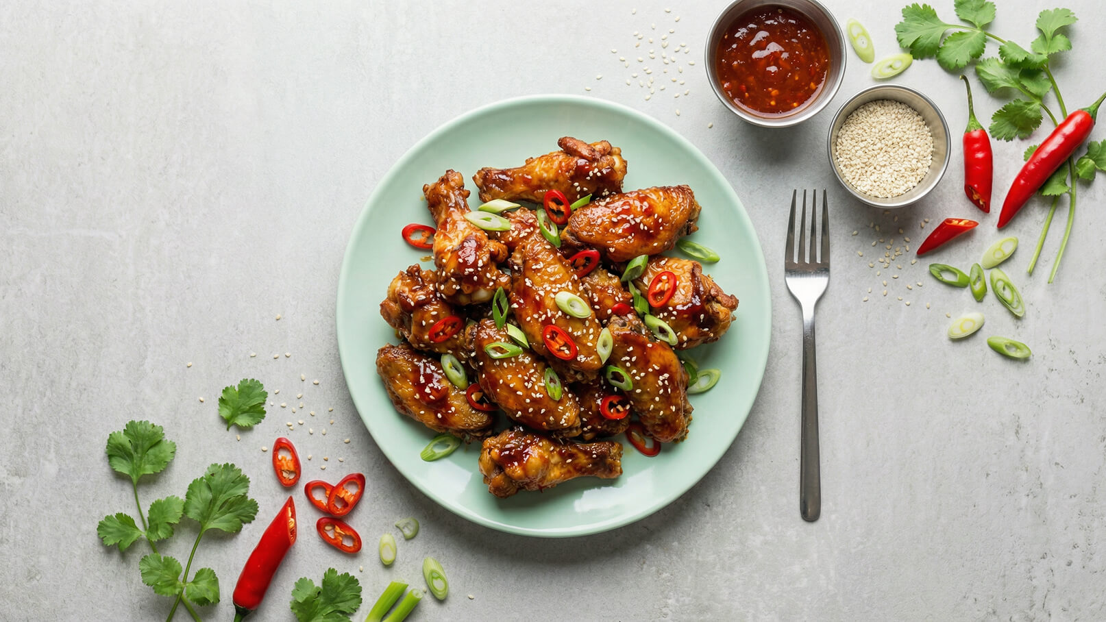 Ein mintgrüner Teller mit knusprigen Hähnchenflügeln, garniert mit gehackten Frühlingszwiebeln und roten Chilischoten. Daneben befinden sich eine Schüssel mit scharfer Sauce, geröstete Sesamsamen und frische Kräuter. Ein Besteck liegt ebenfalls auf dem Tisch.