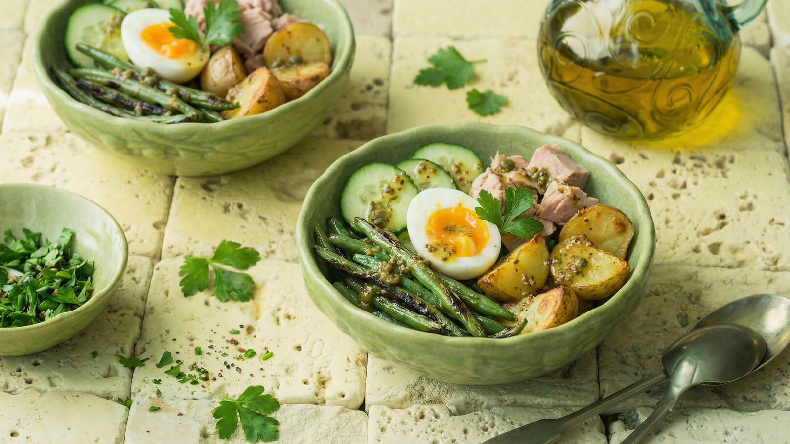 Zwei grüne Schalen mit einer bunten Mischung aus gekochten Eiern, Thunfisch, Kartoffeln, Gurken, grünen Bohnen sowie einer Kräutersauce. Frische Petersilie und ein kleiner Behälter mit gehackten Kräutern befinden sich im Vordergrund. Im Hintergrund ist eine Glasflasche mit Öl.
