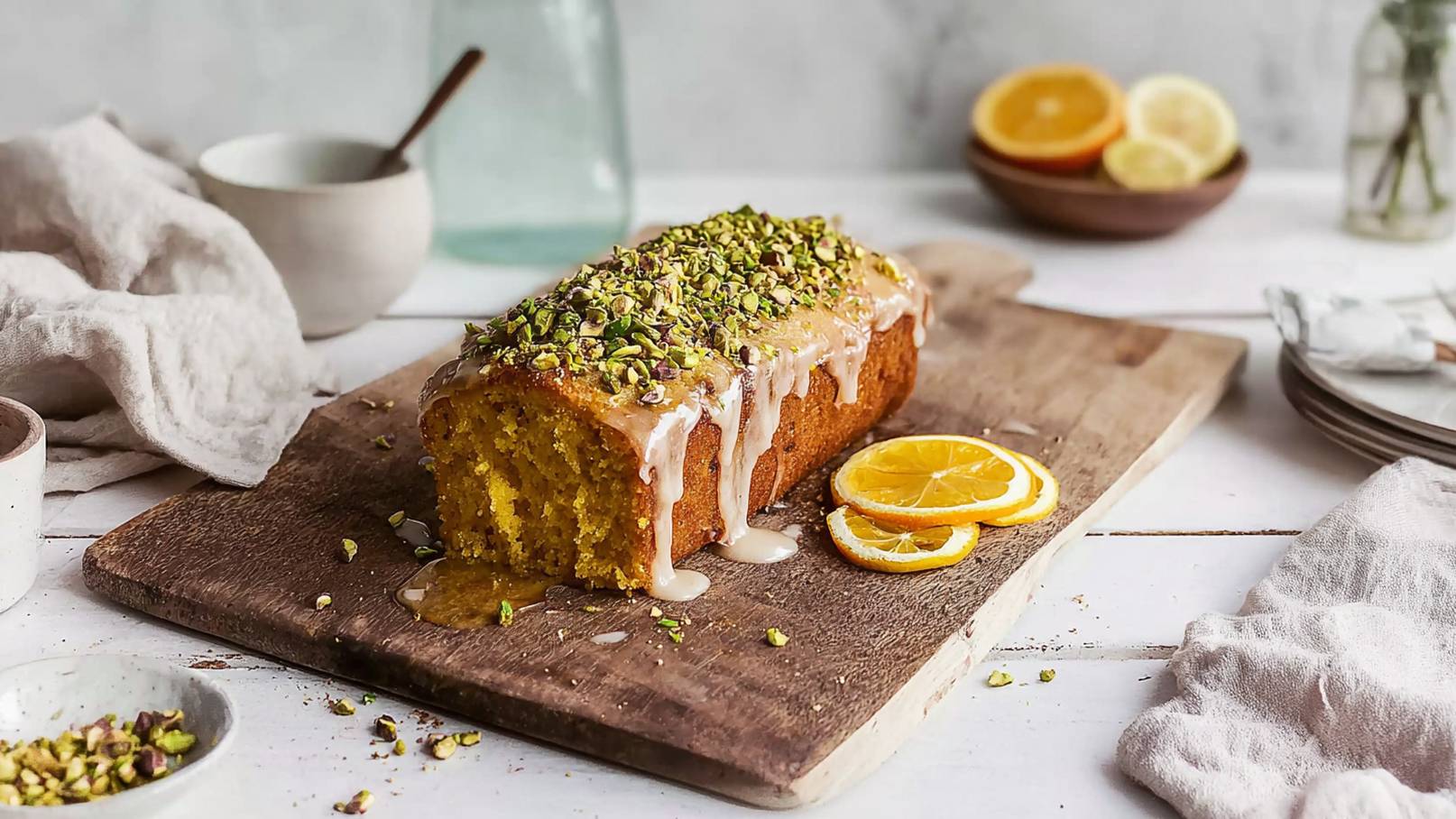 Ein saftiger, mit Zuckerguss überzogener Zitronenkuchen liegt auf einem Holzbrett. Die Oberfläche ist mit gehackten Pistazien dekoriert. Neben dem Kuchen befinden sich frische Zitronenscheiben. Der Hintergrund ist hell und schlicht, wodurch der Kuchen im Vordergrund steht.