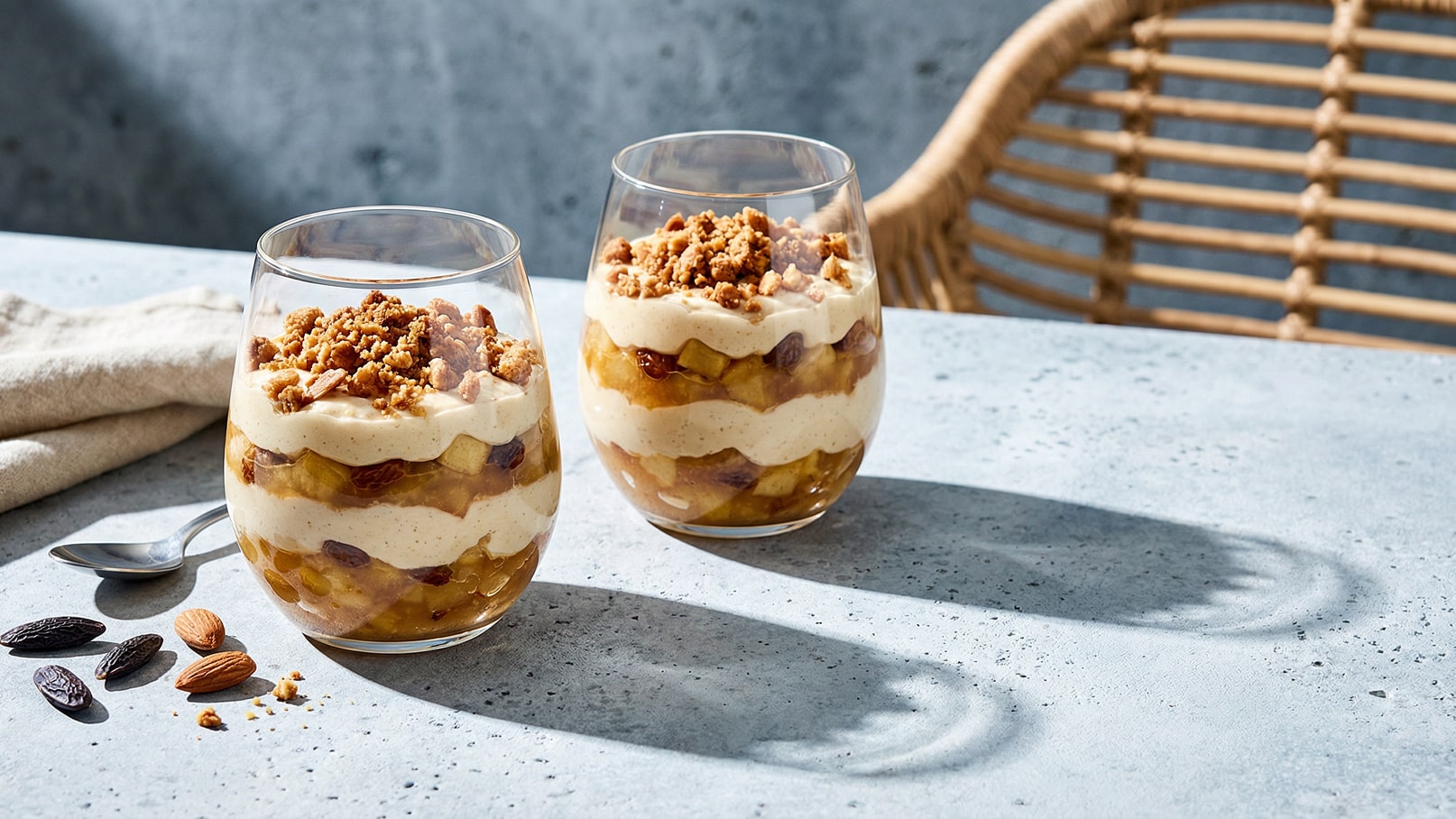 Zwei Gläser mit geschichtetem Dessert sind auf einem Tisch platziert. Die Schichten bestehen aus cremigem Pudding, Äpfeln und Rosinen, gekrönt mit einem knusprigen Kekscrumb. Neben den Gläsern liegen einige Mandeln und ein Löffel auf einer Tischdecke.