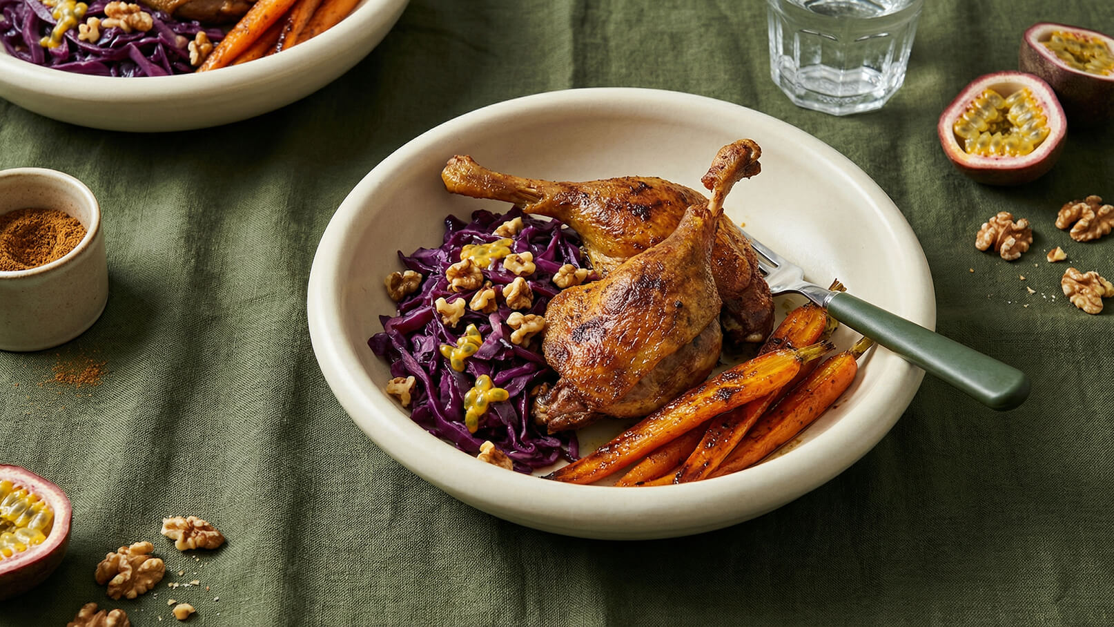 Ein Teller mit gebratenem Hähnchenschenkel, roten Kohl, Walnüssen und gerösteten Karotten. Die Speisen sind auf einem beigefarbenen Teller angerichtet. Im Hintergrund befindet sich ein Glas Wasser und eine Schale mit Gewürzen. Der Tisch ist mit einem grünen Tuch bedeckt.