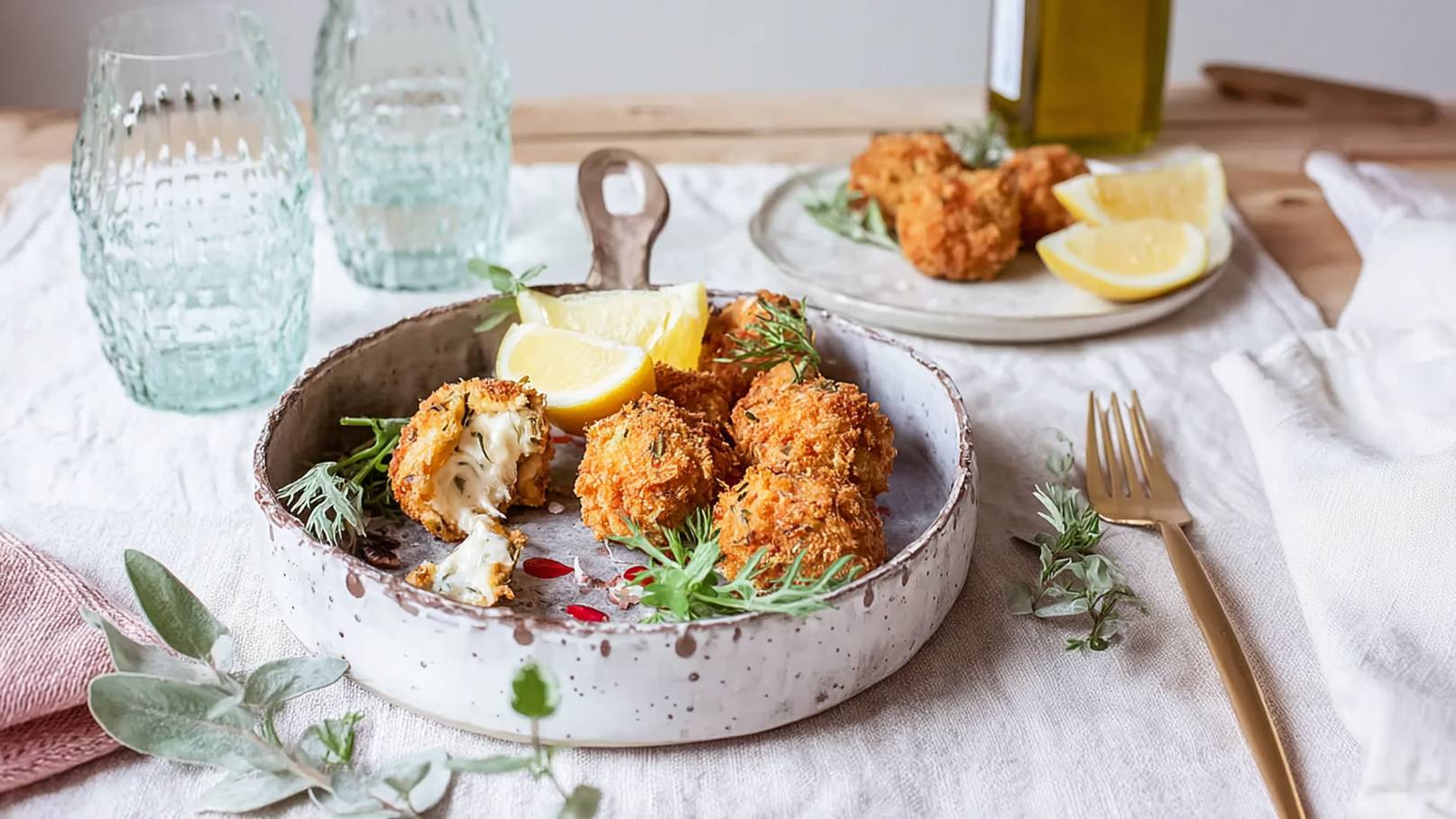Ein rustikeller Teller mit knusprigen, goldbraunen Bällchen, die leicht geöffnet sind und Käse zeigen. Dazu sind frische Zitronenscheiben und grüne Kräuter auf dem Teller dekoriert. Im Hintergrund sind zwei Gläser und ein Olivenölbehälter sichtbar.