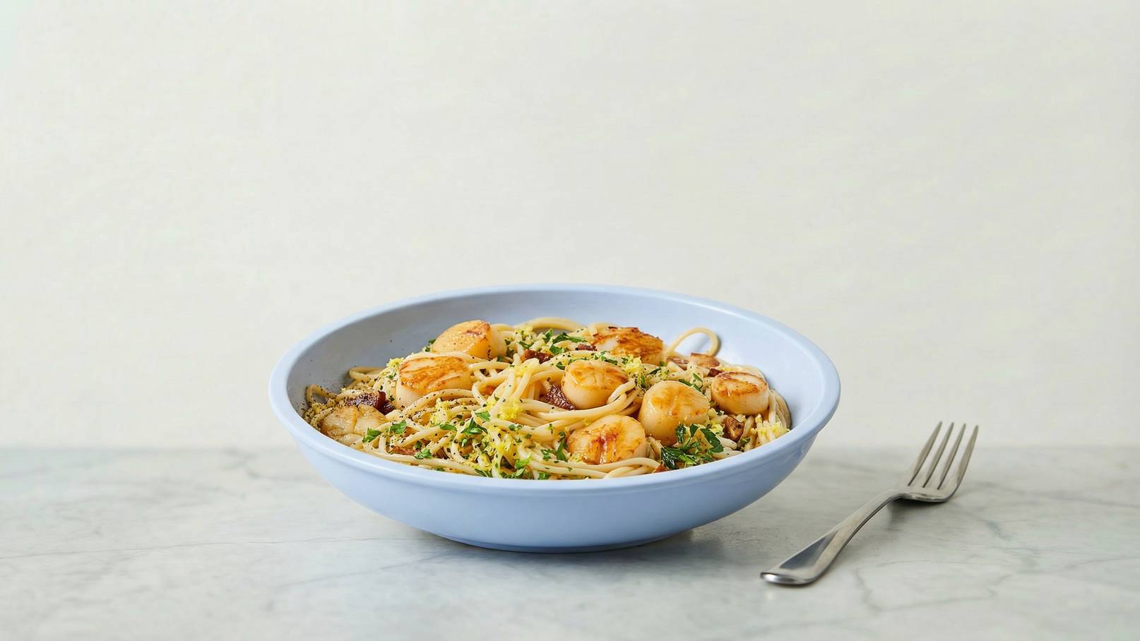 Ein blauer Teller mit Pasta, garniert mit gebratenen Jakobsmuscheln und frischen Kräutern. Die Nudeln sind gut sichtbar, und die goldbraunen Muscheln sind gleichmäßig verteilt. Im Hintergrund liegt ein Besteckstück.