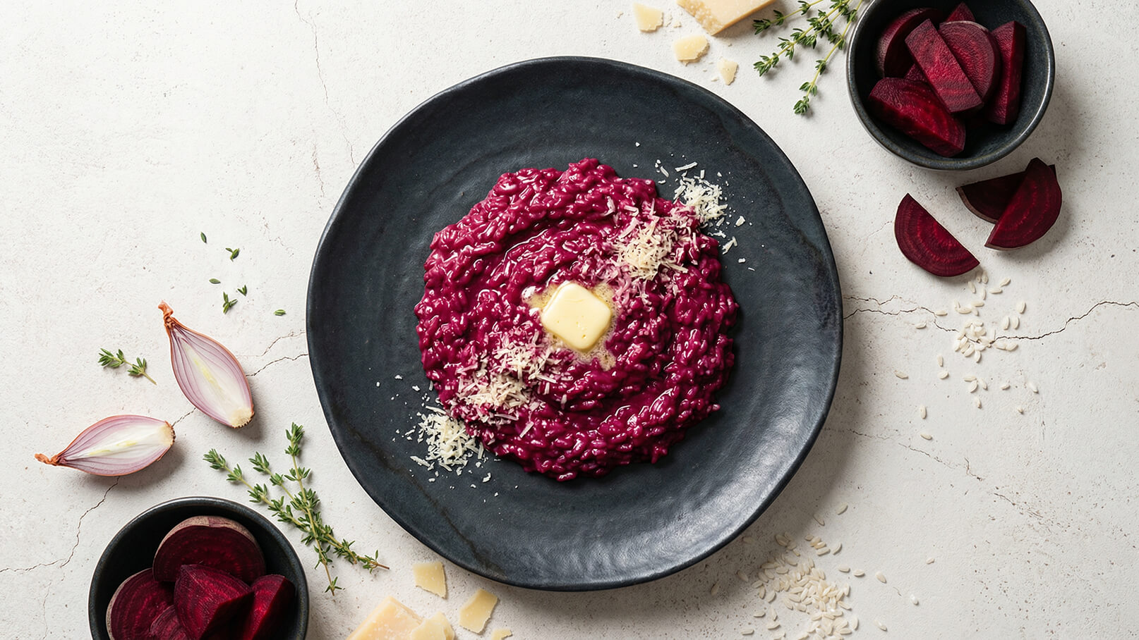 Ein schwarzer Teller enthält lila Risotto, das mit Butter und geriebenem Käse garniert ist. Daneben liegen frische Rote Bete und einige Kräuter. Die Präsentation ist elegant und ansprechend, ideal für ein feines Gericht.