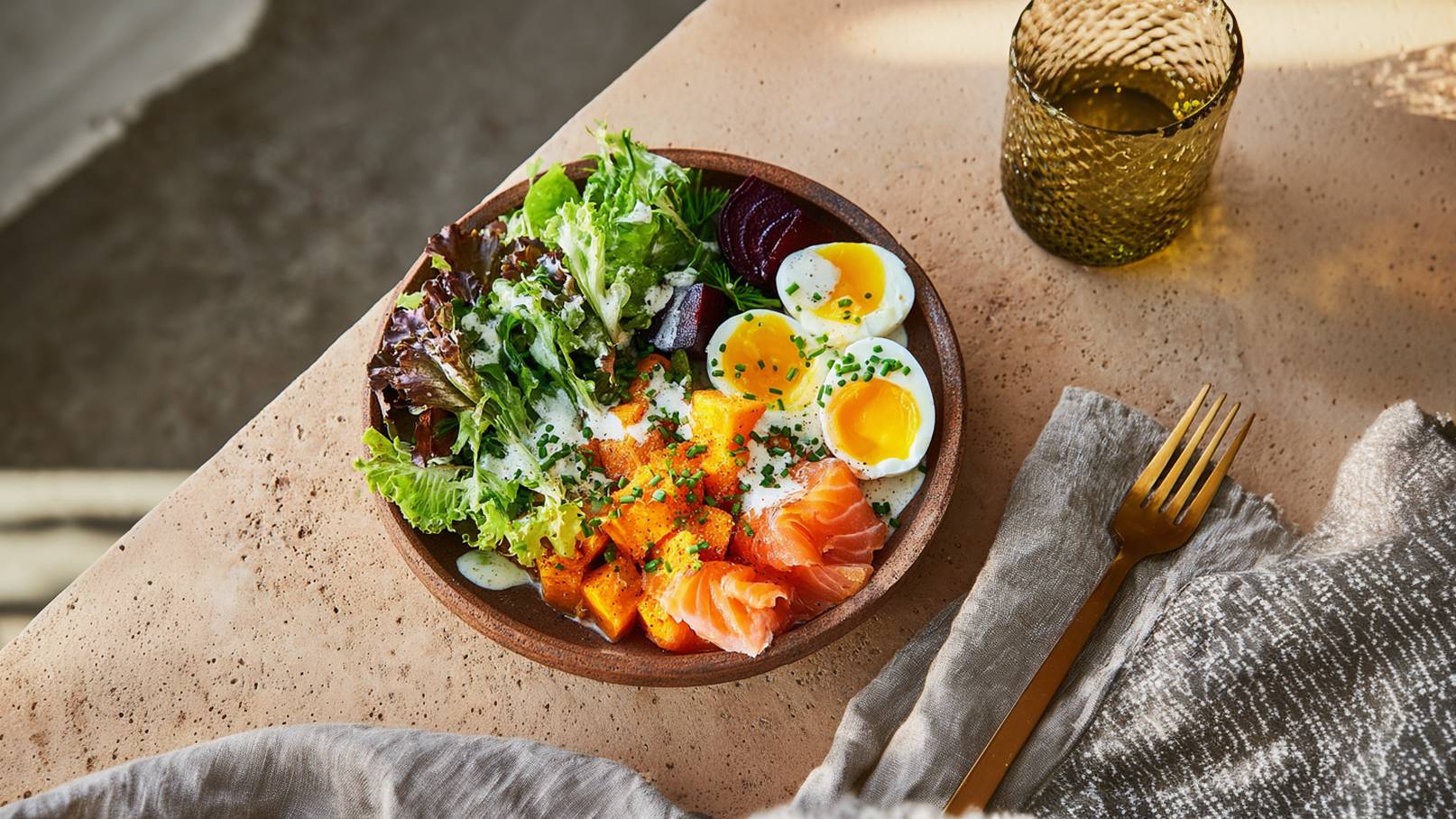 Ein rustikeller Teller mit frischem Salat, Räucherlachs, gekochten Eiern und Kürbisstücken. Das Gericht ist garnieren mit Schnittlauch und einem cremigen Dressing. Im Hintergrund befindet sich ein dekoratives Glas und eine goldene Gabel auf einem Tisch.