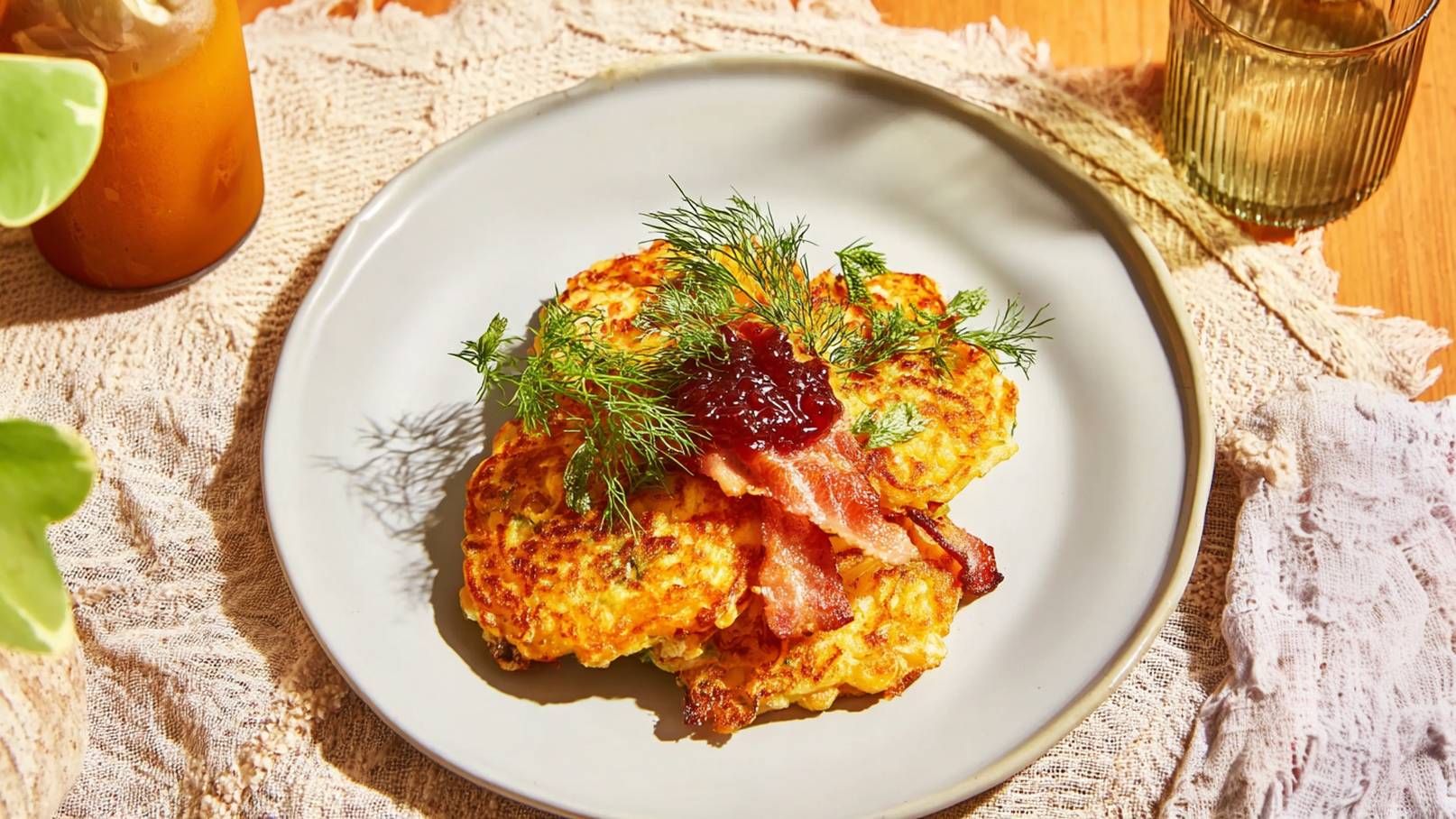 Ein Teller mit gebratenen Kürbis-Pfannkuchen, garniert mit frischem Dill, knusprigem Bacon und einem Klecks Marmelade. Der Teller steht auf einer hellen Tischdecke, umgeben von einem Getränk und weiteren Tischaccessoires.
