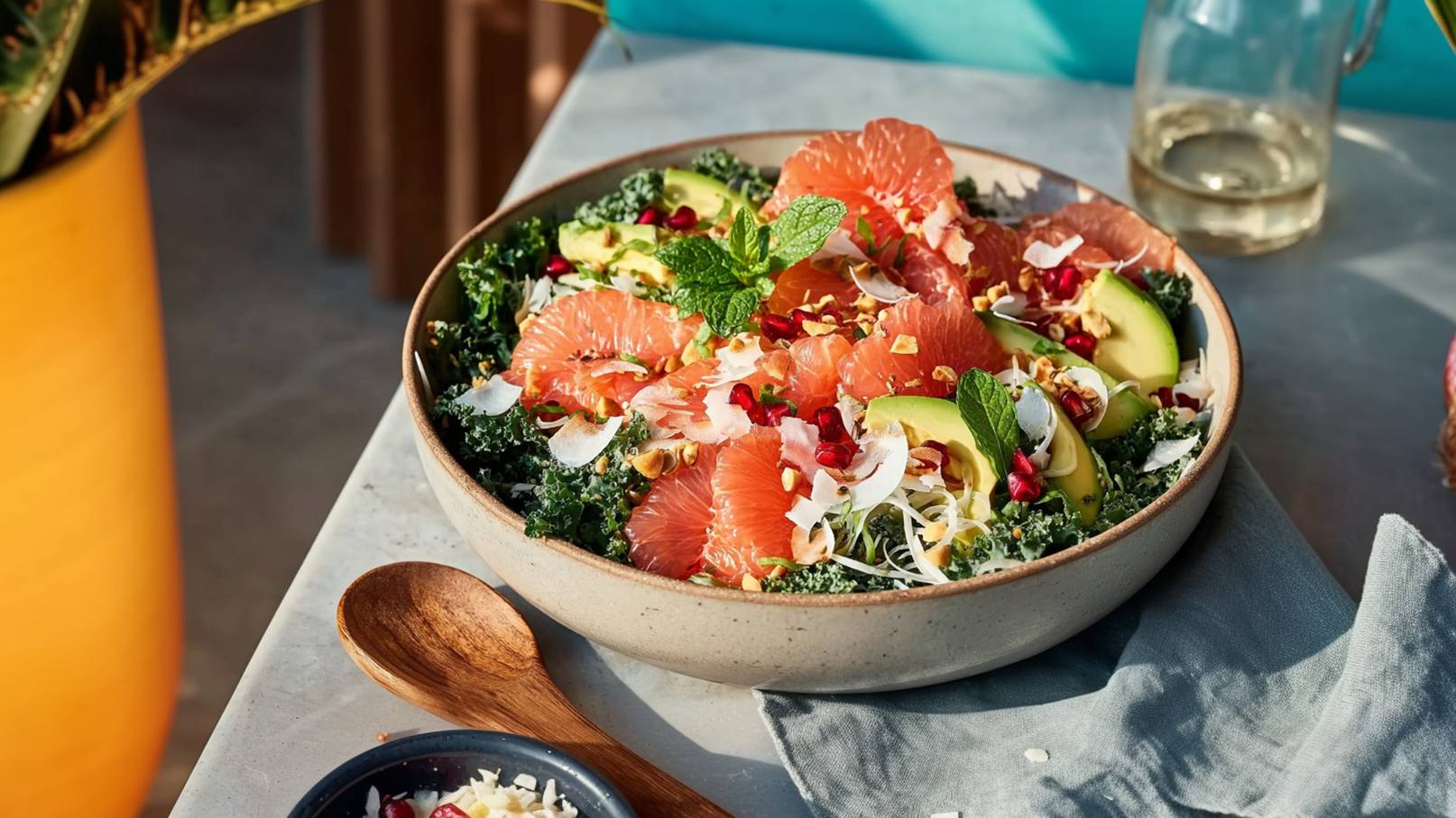 Eine Schüssel mit grünem Blattgemüse, belegt mit rosa Grapefruit, Avocado, Minzblättern, Granatapfelkernen und gerösteten Nüssen. Der Salat ist ansprechend angerichtet und wird auf einem hellen Tisch präsentiert. Daneben liegt ein Holzlöffel und ein Stofftuch.