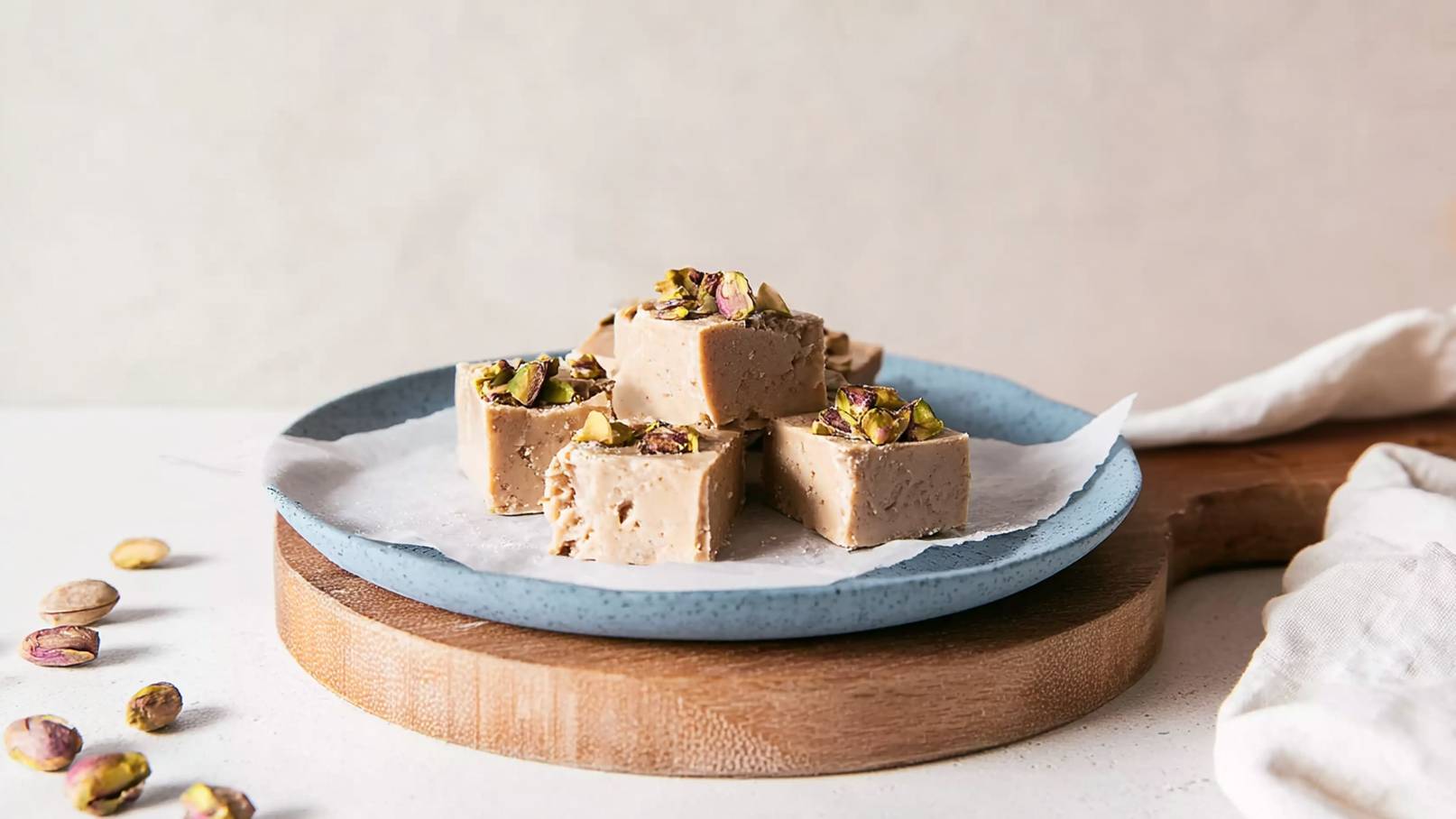 Eine blaue Platte mit einer haufartigen Anordnung von quadratischen, hellbraunen Stückchen, die mit gehackten Pistazien dekoriert sind. Die Süßigkeit liegt auf einem Blatt Backpapier. Um die Platte herum sind einige Pistaziensamen verteilt.