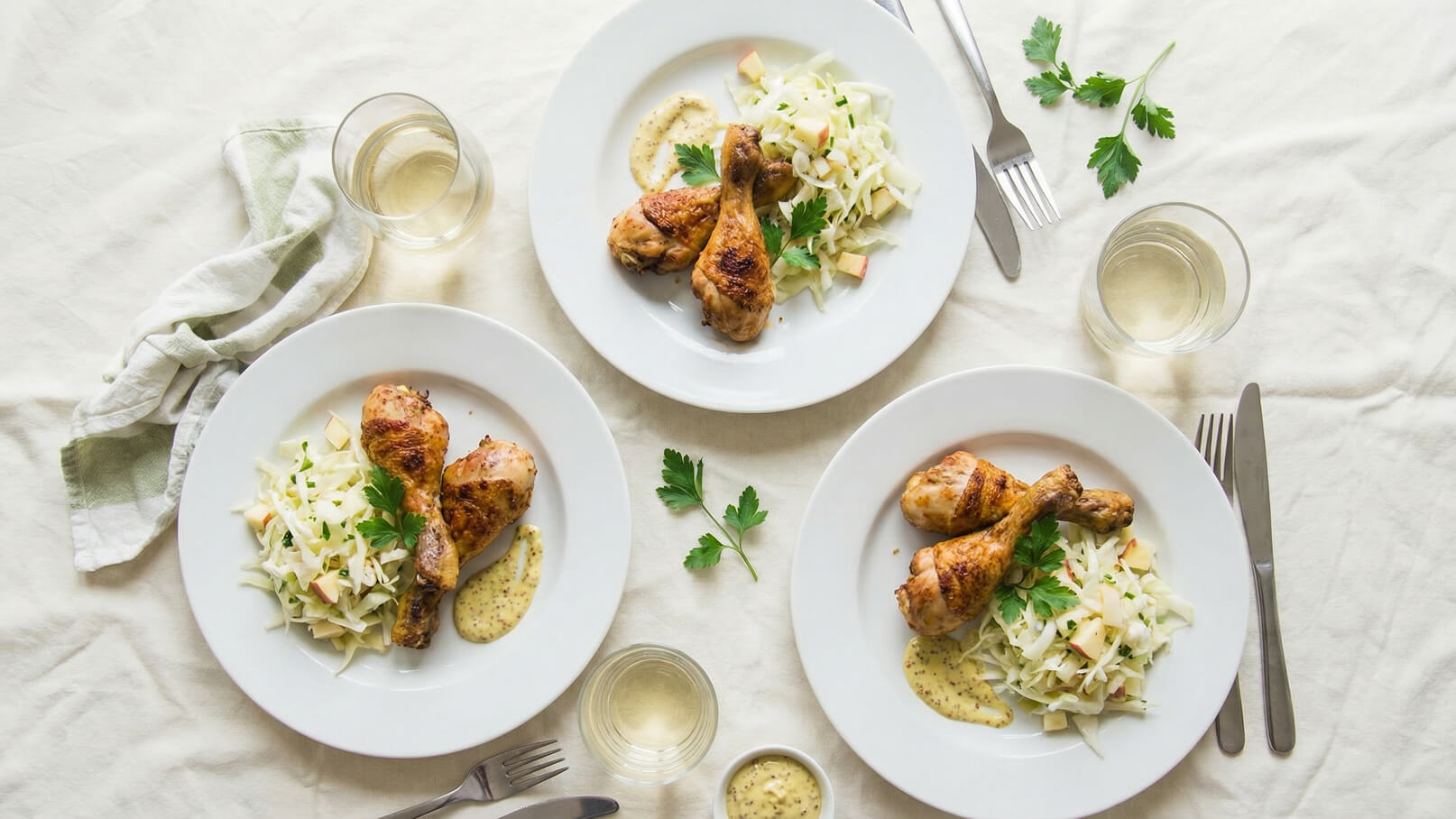 Drei Teller mit gegrillten Hähnchenschenkeln, umgeben von einem Apfel-Kohl-Salat und Senfsoße. Ein Glas Wasser steht neben den Tellern auf einem hellen Tisch. Frische Petersilie dekoriert die Teller.