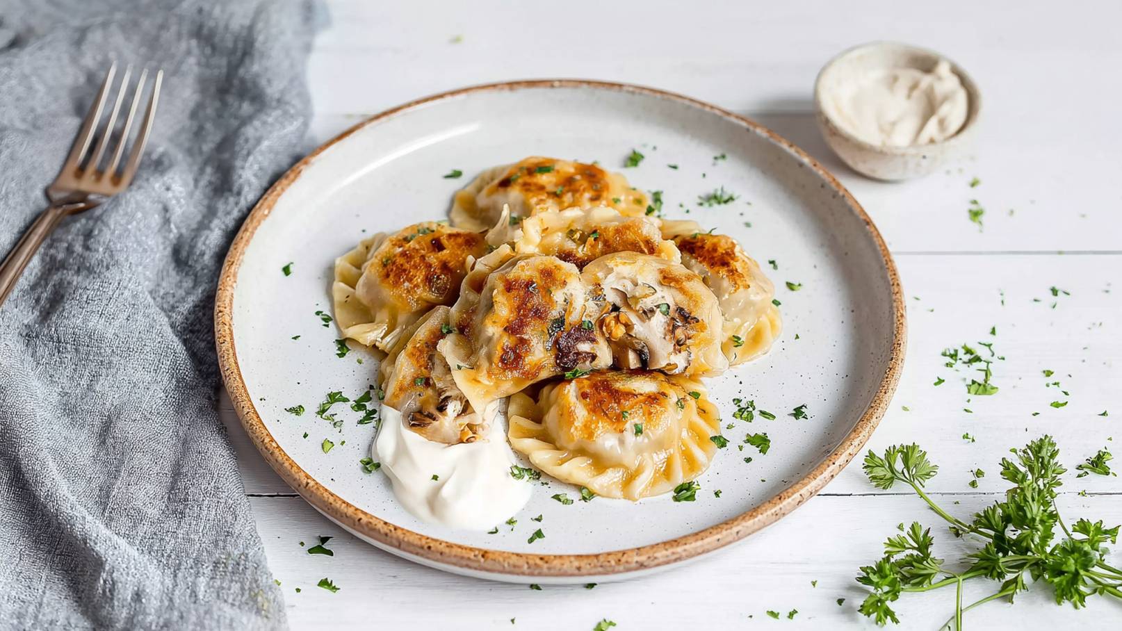 Ein Teller mit golden gebratenen Teigtaschen, gefüllt mit Pilzen und Kräutern. Die Teigtaschen liegen auf einem weißen Teller, umgeben von frischer Petersilie. Neben dem Teller befindet sich ein kleiner Schälchen mit Sauerrahm.