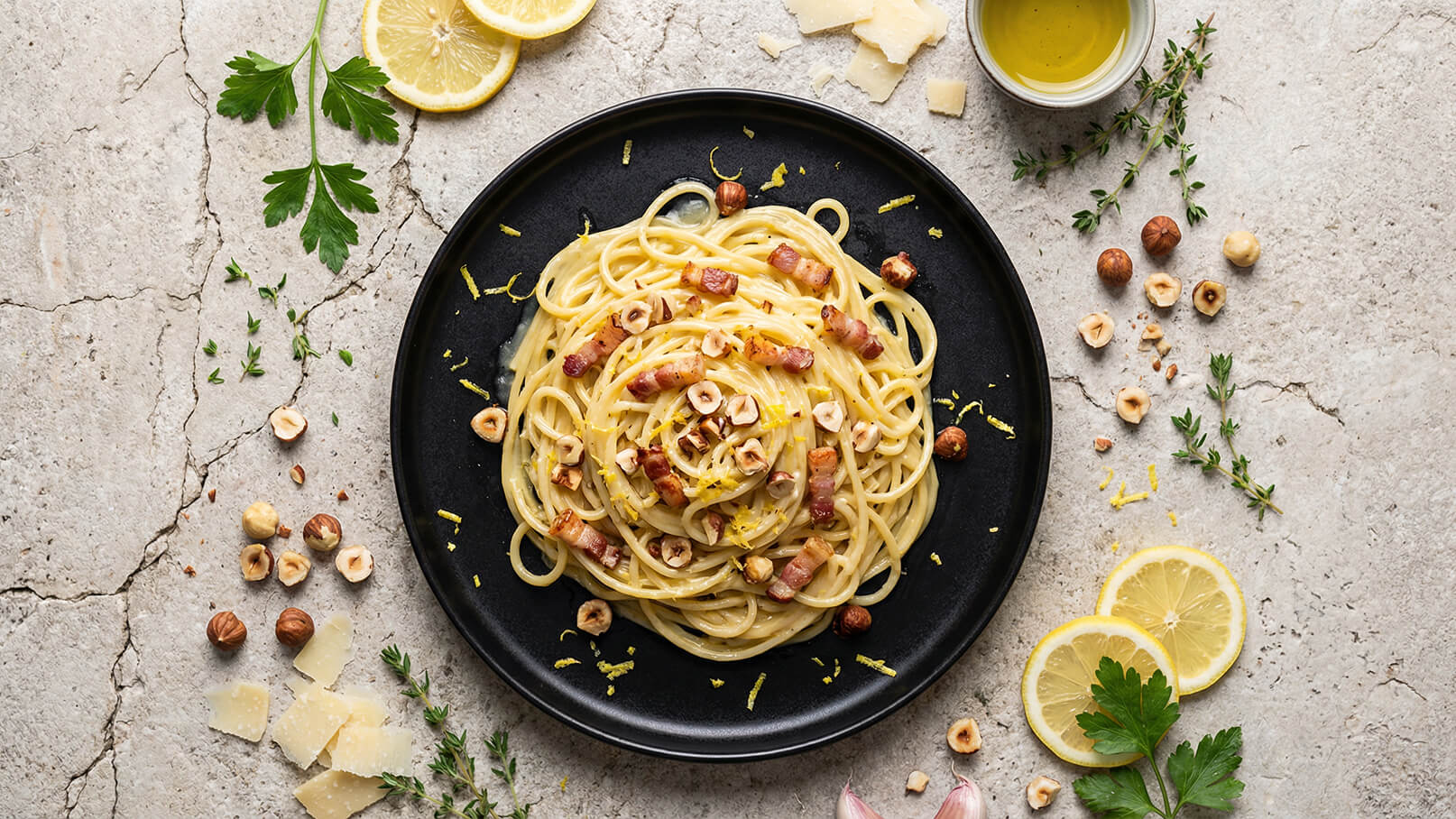Ein schwarzes Teller mit Spaghetti, die mit Stückchen von Speck und gehackten Haselnüssen garniert sind. Darauf liegt Zitronenschale. Um das Teller herum sind Zitronenscheiben, frische Kräuter und Parmesanstücke angeordnet.
