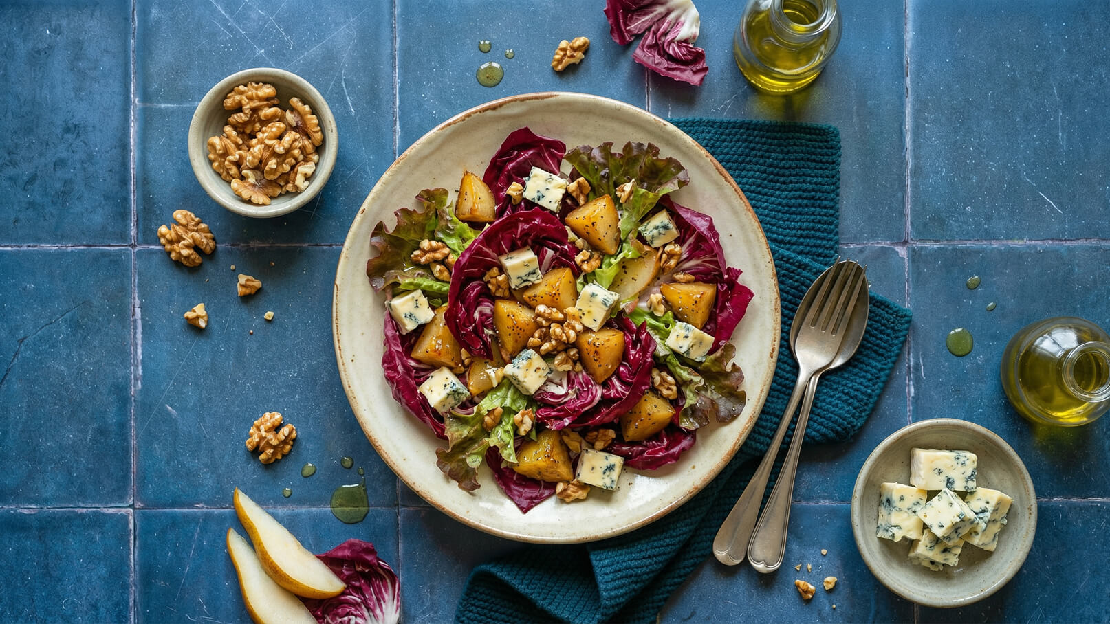 Ein Teller mit buntem Salat, bestehend aus rotgrünem Blattgemüse, stückigen Birnen, Walnüssen und Käsestücken. Daneben eine kleine Schale mit Walnüssen und ein Tee-Handtuch, sowie ein paar Esslöffel aus Metall. Die Dekoration ist schlicht und ansprechend.