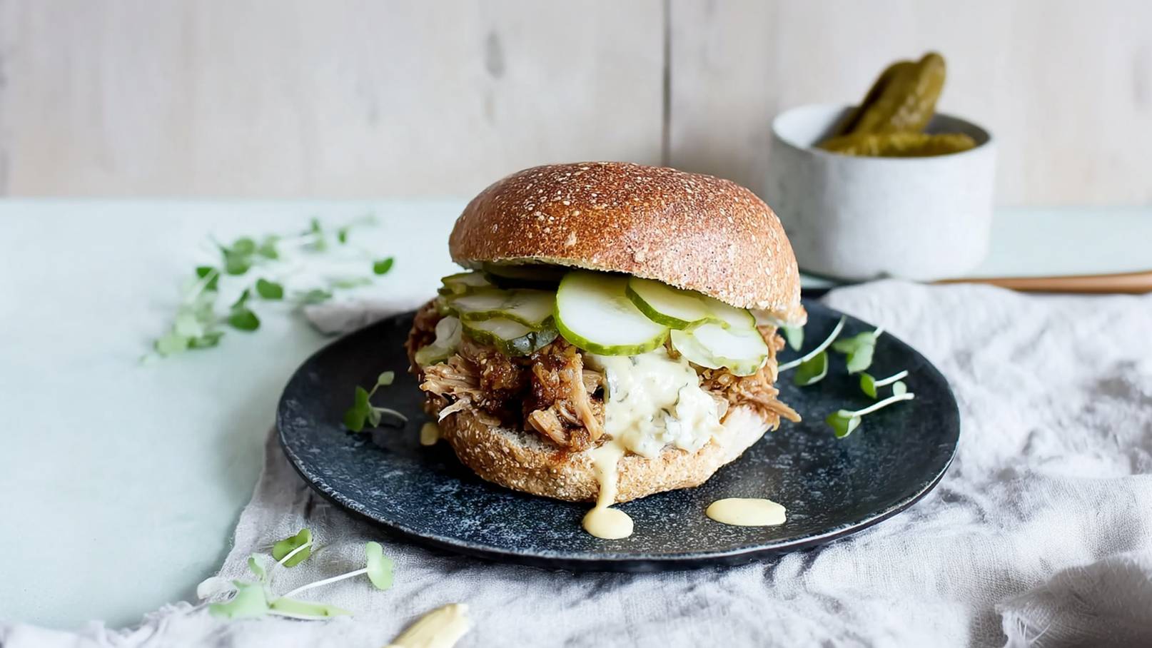 Ein frisch zubereiteter Burger auf einem schwarzen Teller. Der Burger besteht aus einem Vollkornbrötchen, gefüllt mit zartem, pulled Fleisch, Saucen und knackigen Gurkenscheiben. Im Hintergrund sind einige grüne Kräuter und ein Glas mit Essiggurken sichtbar.