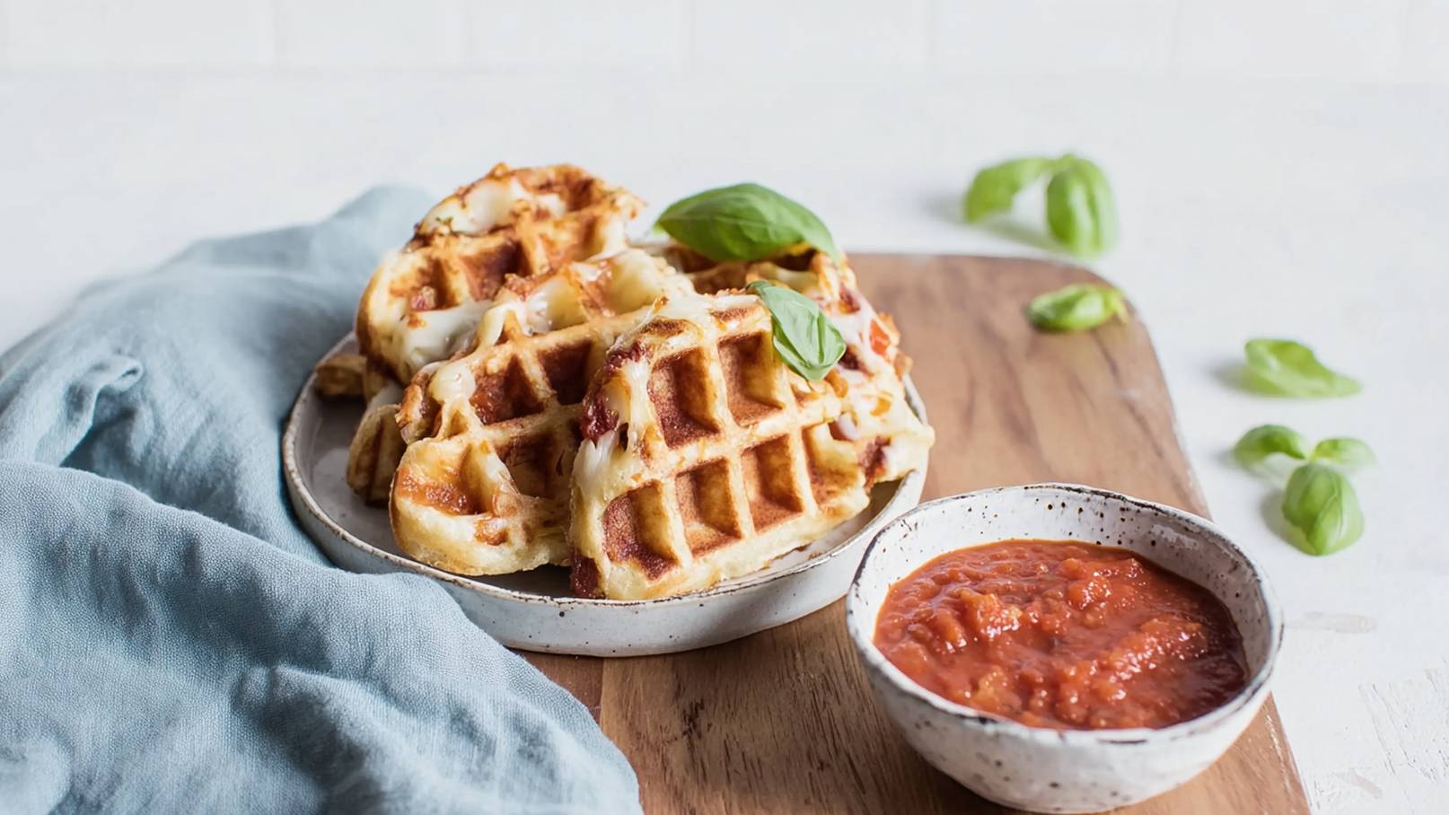 Auf einem Holzteller liegen mehrere goldbraune Waffeln zusammen mit einer kleinen Schüssel Tomatensoße. Einige frische Basilikumblätter sind um die Waffeln und die Schüssel verteilt, was dem Bild einen frischen Touch verleiht.