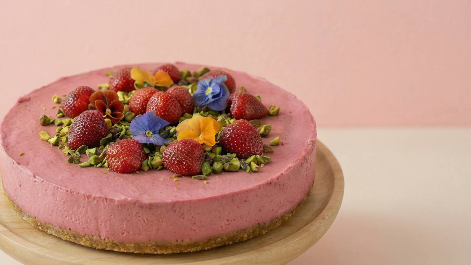 Eine runde Erdbeertorte auf einem Holzuntergrund, die mit frischen Erdbeeren, bunten Blüten und gehackten Pistazien dekoriert ist. Die Torte hat eine zarte rosa Farbe und wirkt ansprechend und festlich. Der Hintergrund ist in sanften Rosa-Tönen gehalten.